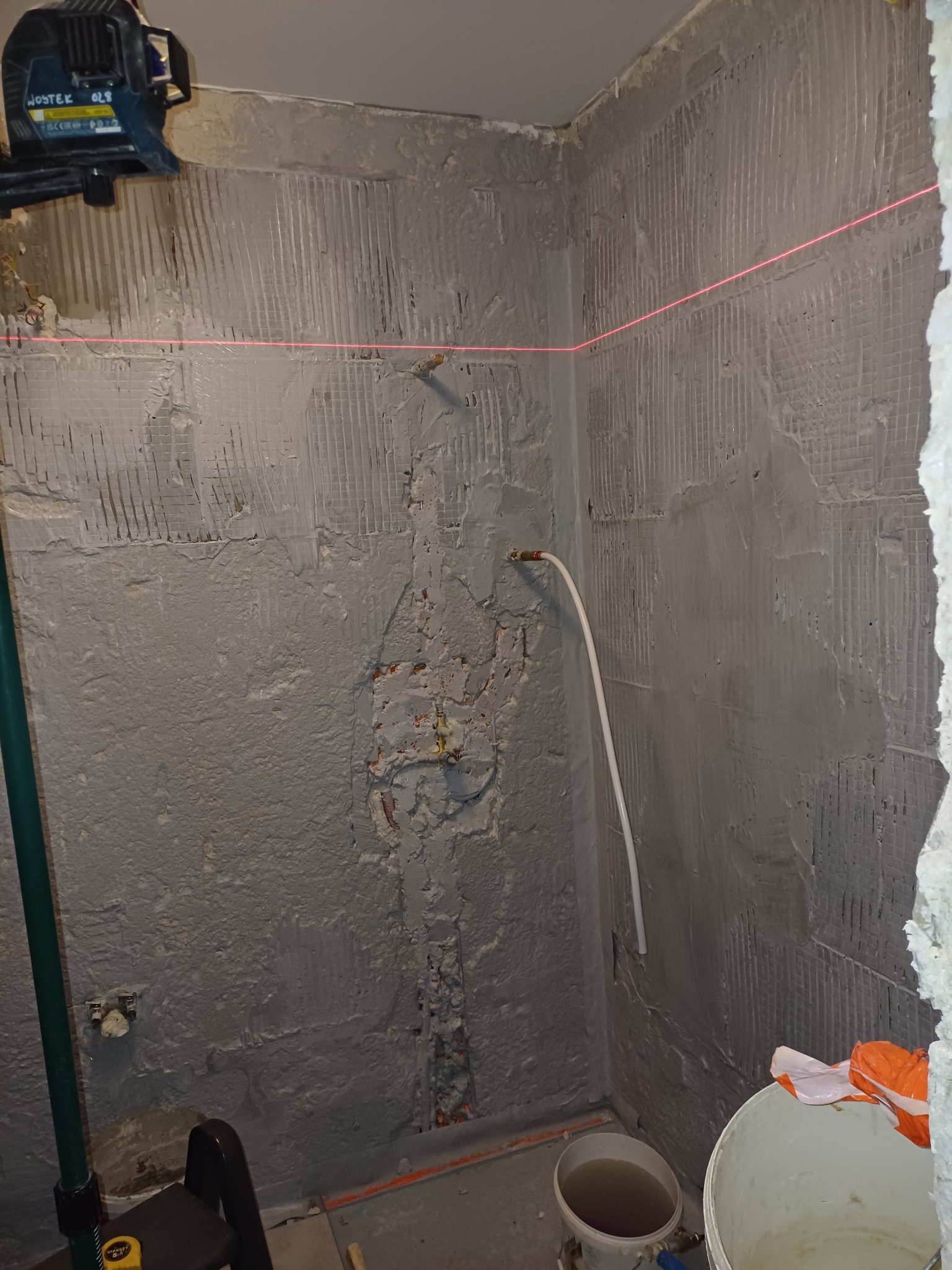 Przygotowanie ściany pod płytki w rogu pomieszczenia, widoczna siatka pod tynkiem, poziomica laserowa wyznacza linię, fragment instalacji wodnej, dwa wiadra na podłodze.