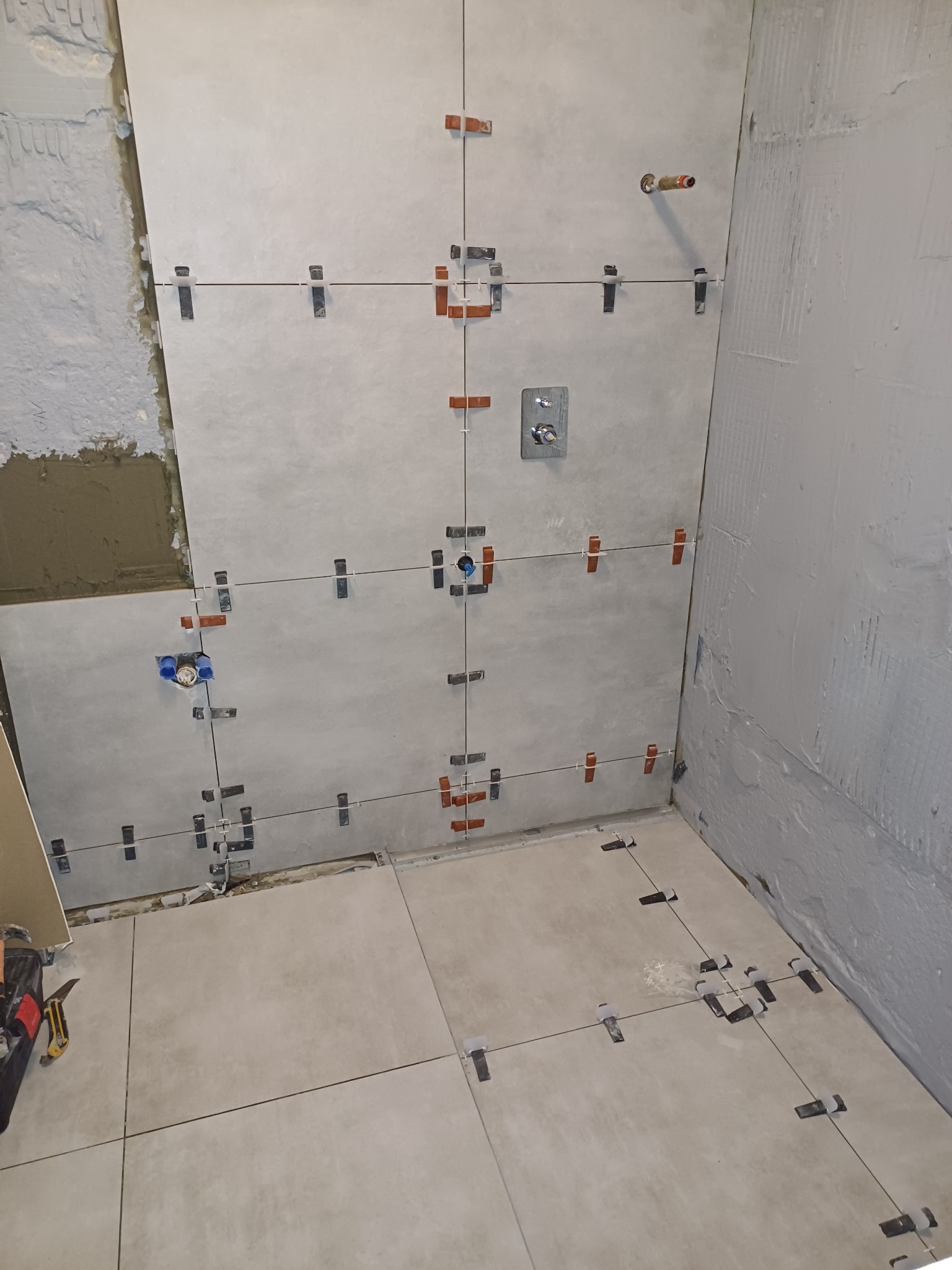 Świeżo ułożone, jasnoszare płytki w łazience, z widocznymi krzyżykami dystansowymi i elementami instalacji wodnej, częściowo na surowym, nieotynkowanym murze.