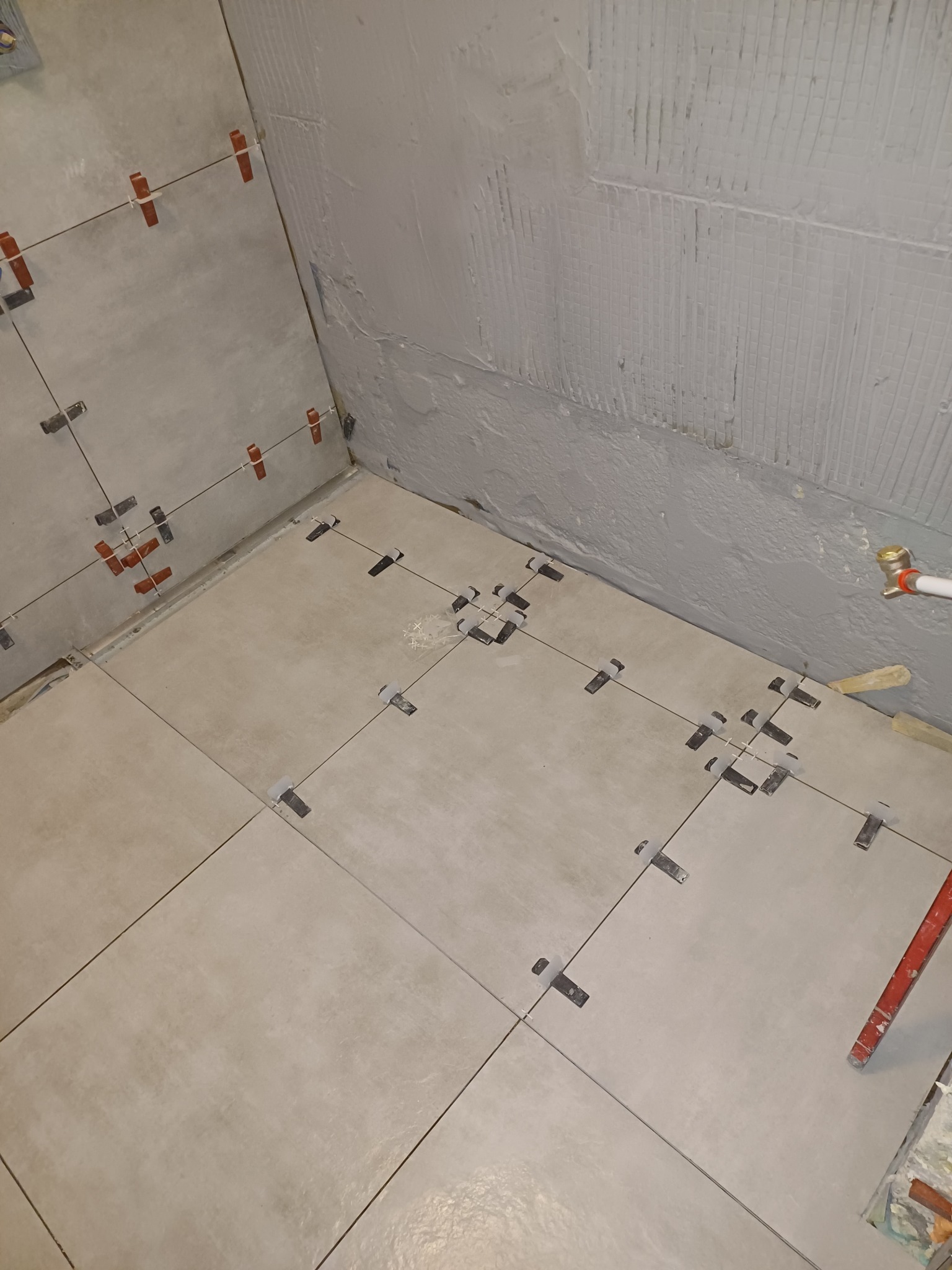 Układanie beżowych płytek podłogowych w rogu pomieszczenia z użyciem klipsów poziomujących i sznurka, świeżo nałożony szary tynk na ścianie w tle.