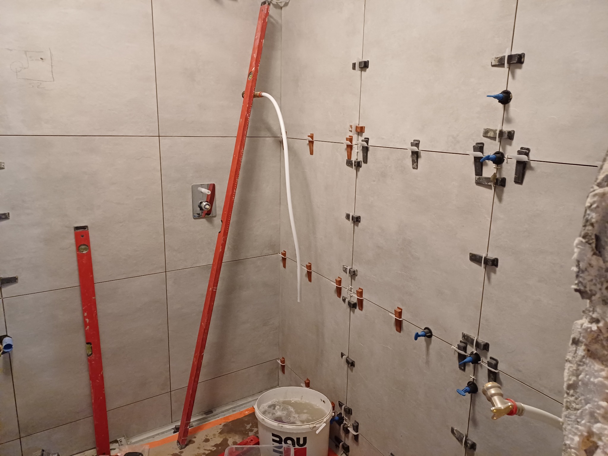 Szare płytki ścienne w trakcie montażu, widoczne poziomice, wiadro z wodą i system poziomowania płytek z niebieskimi elementami, fragment instalacji wodnej.