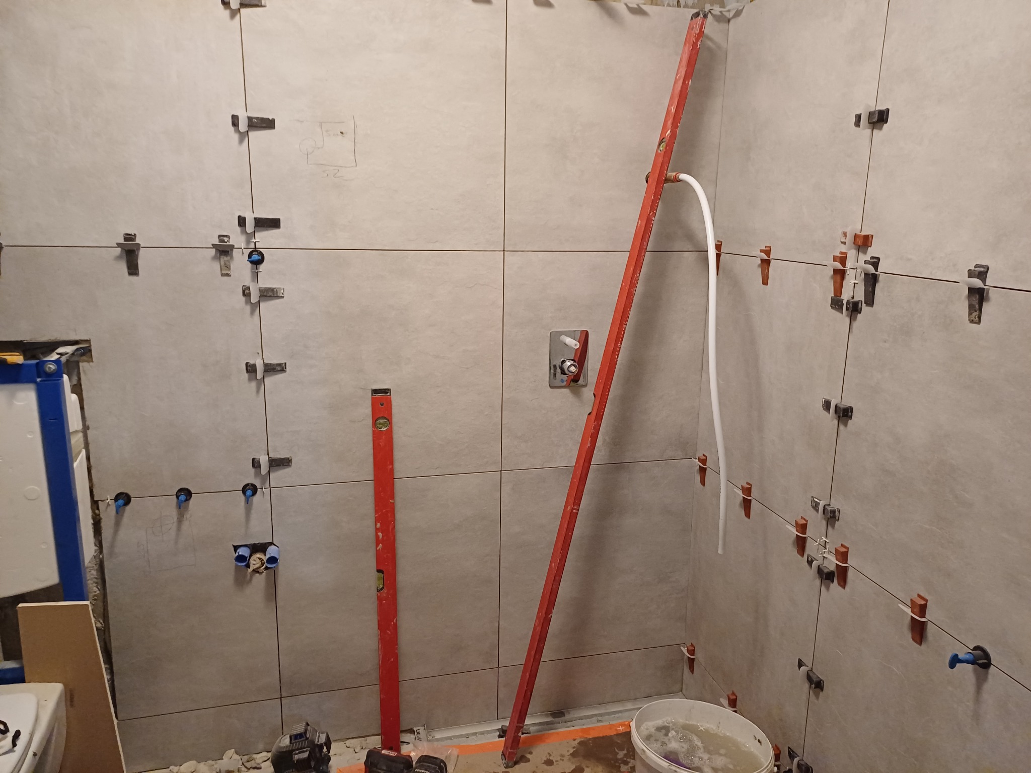 Ściana w trakcie układania płytek, widoczne poziomice, krzyżyki dystansowe, rury i przygotowane otwory na armaturę.