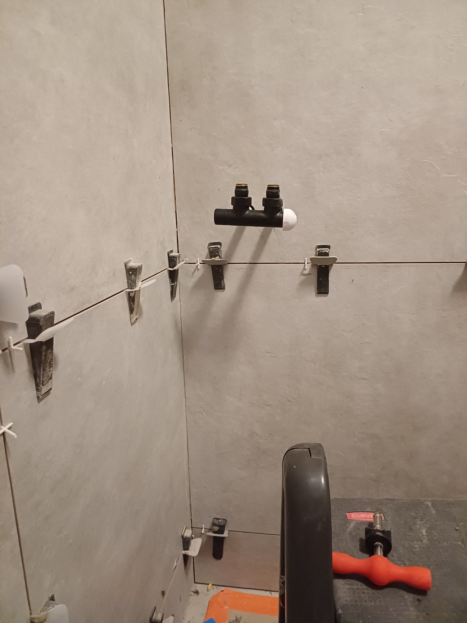 Ściana wyłożona szarymi płytkami, z widocznymi systemami poziomowania płytek i fragmentem instalacji wodnej oraz narzędziem do układania płytek w dolnej części kadru.