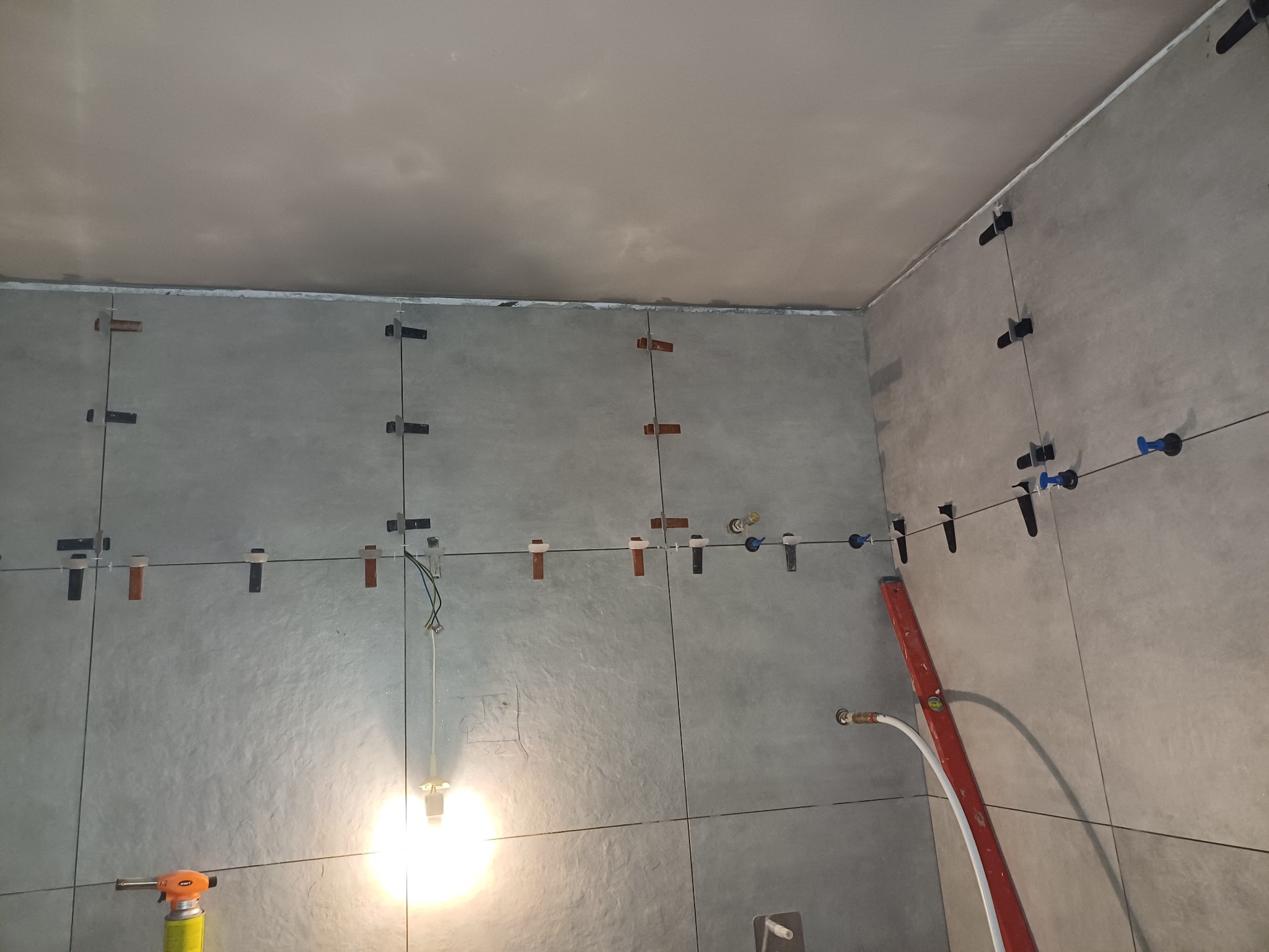 Świeżo ułożone, szare płytki ścienne z systemem poziomowania, widoczne przewody elektryczne i palnik gazowy w rogu pomieszczenia.