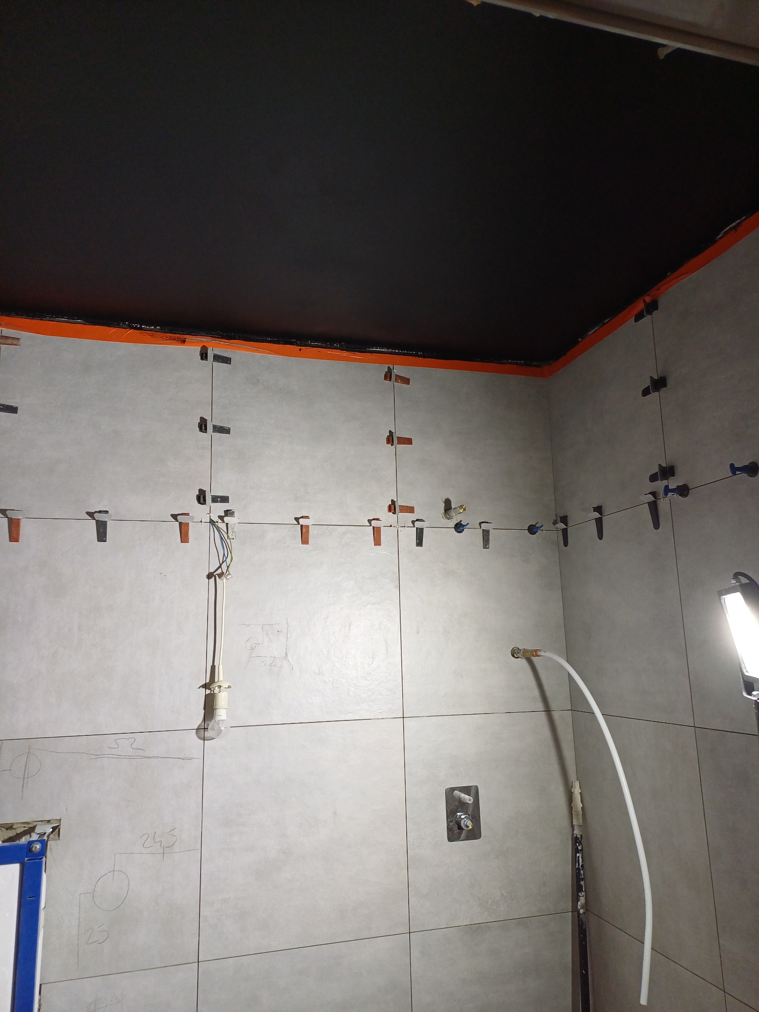 Ściana w trakcie układania płytek ceramicznych, widoczne krzyżyki dystansowe, przewody elektryczne, rury i prowizoryczne oświetlenie, na ścianie widoczne odręczne notatki wymiarowe.