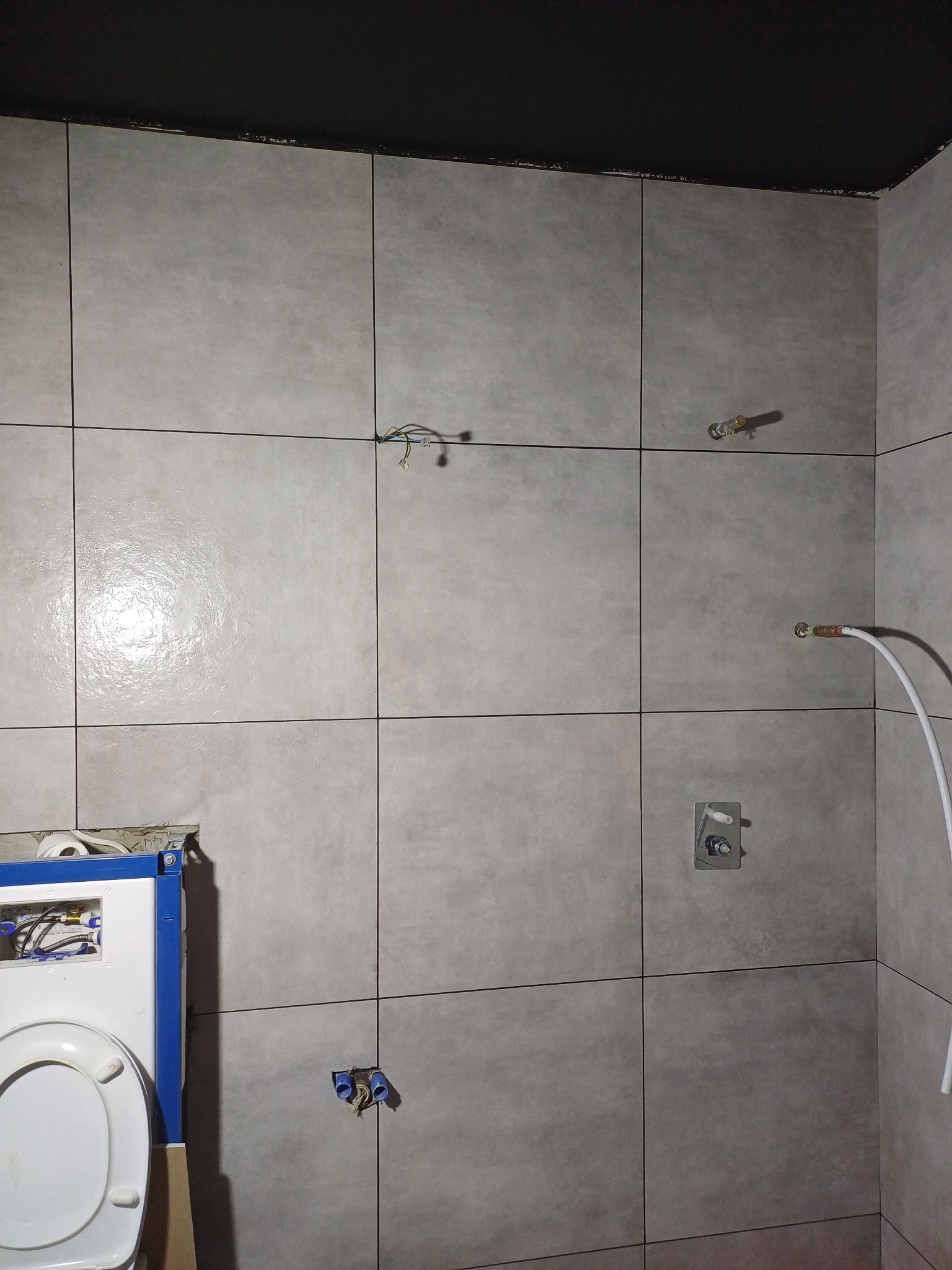 Szare, matowe płytki ścienne w trakcie montażu w łazience, widoczne wystające przewody elektryczne, instalacja wodna i częściowo zamontowana toaleta podtynkowa.