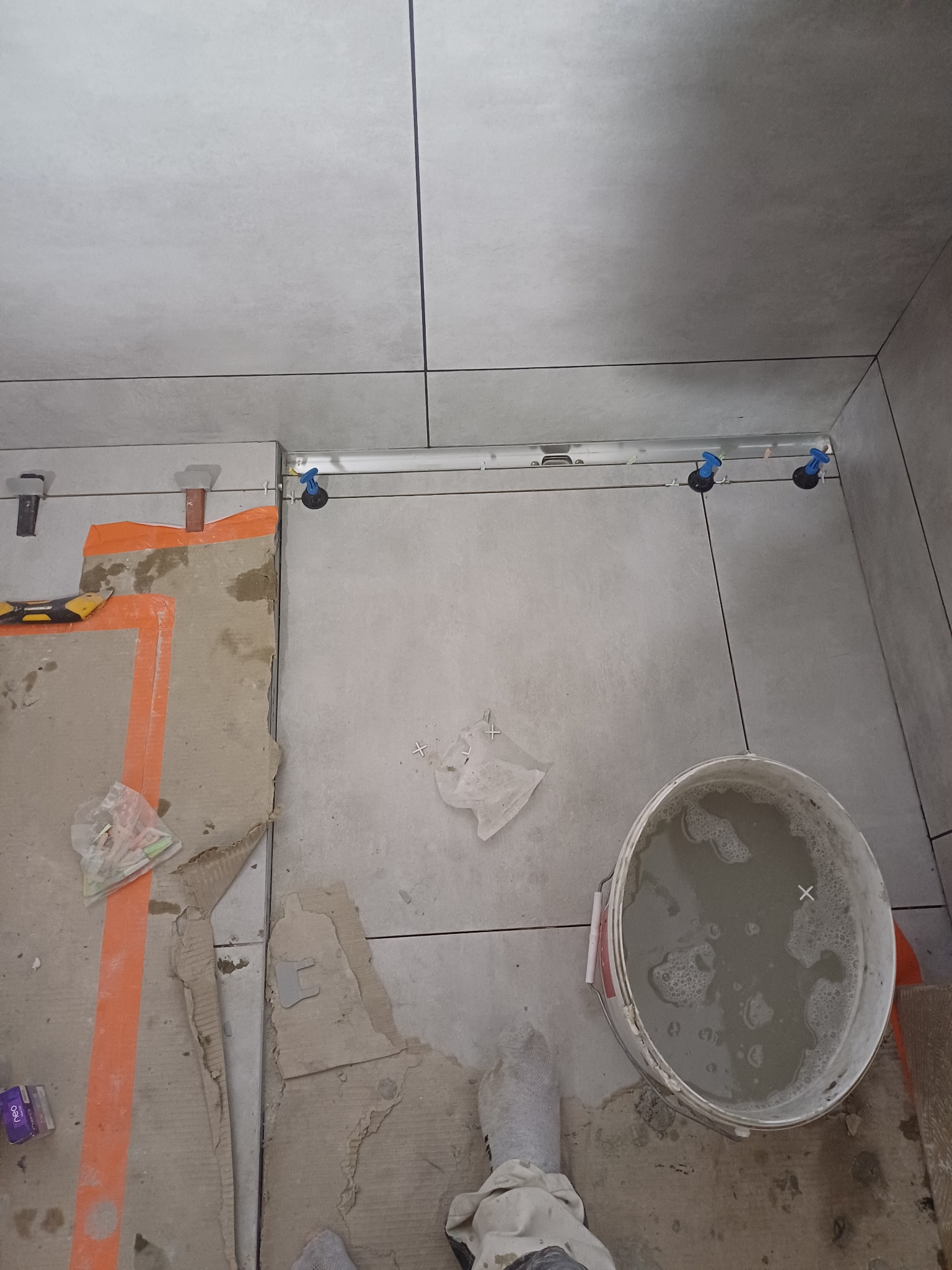 Szare płytki na podłodze, w trakcie układania, widoczne narzędzia poziomujące i wiadro z zaprawą, ochraniacz na but.