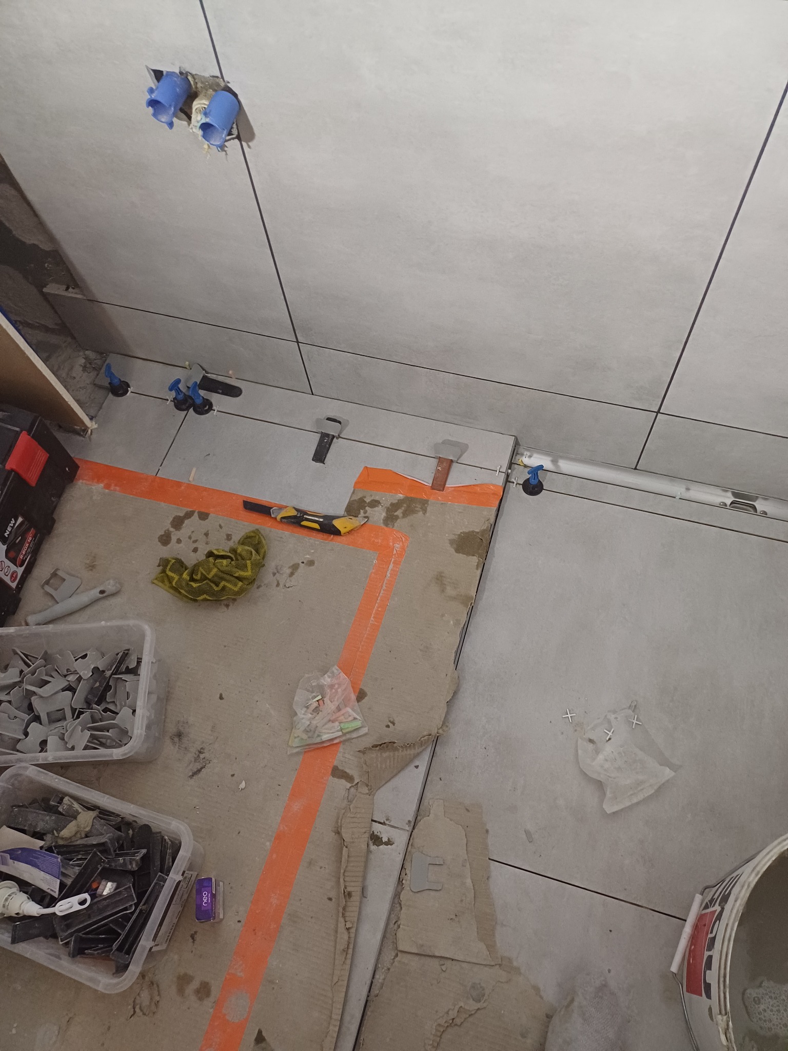 W trakcie układania jasnoszarych płytek ściennych i podłogowych z użyciem systemu poziomowania, widoczne narzędzia, poziomica, pojemniki z klipsami i klinami, oraz taśma malarska wyznaczająca...