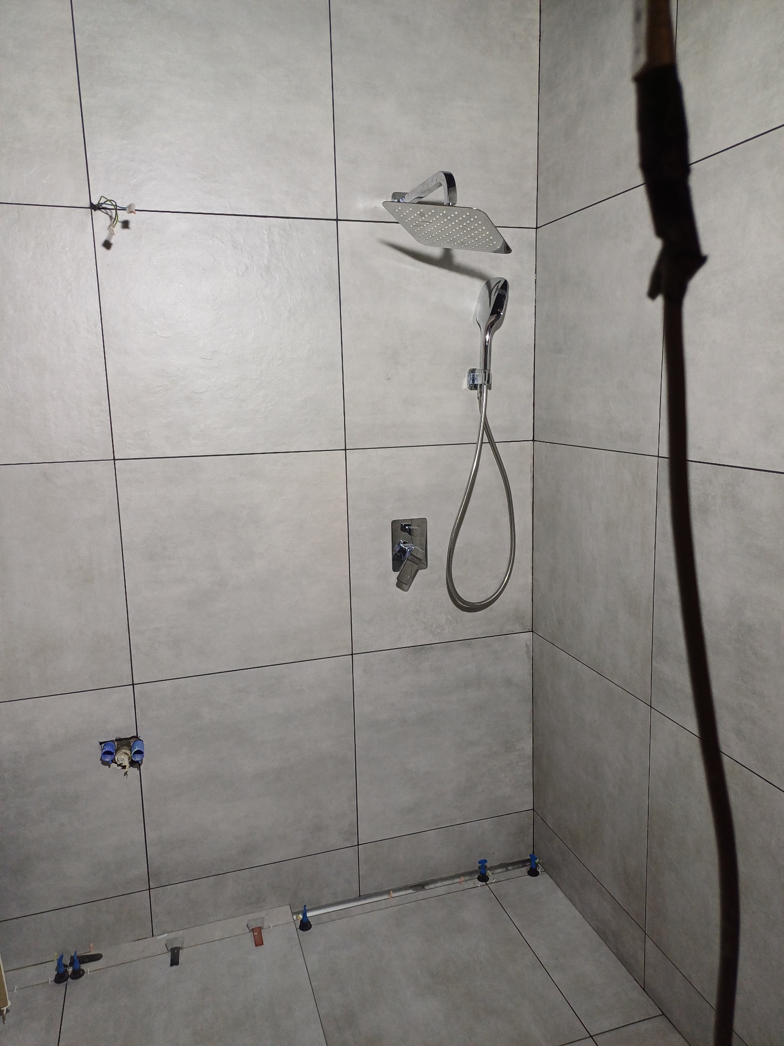 Szare płytki w łazience, zamontowana deszczownica i bateria, widoczne elementy instalacji wodnej i poziomica z niebieskimi klinami do układania płytek.