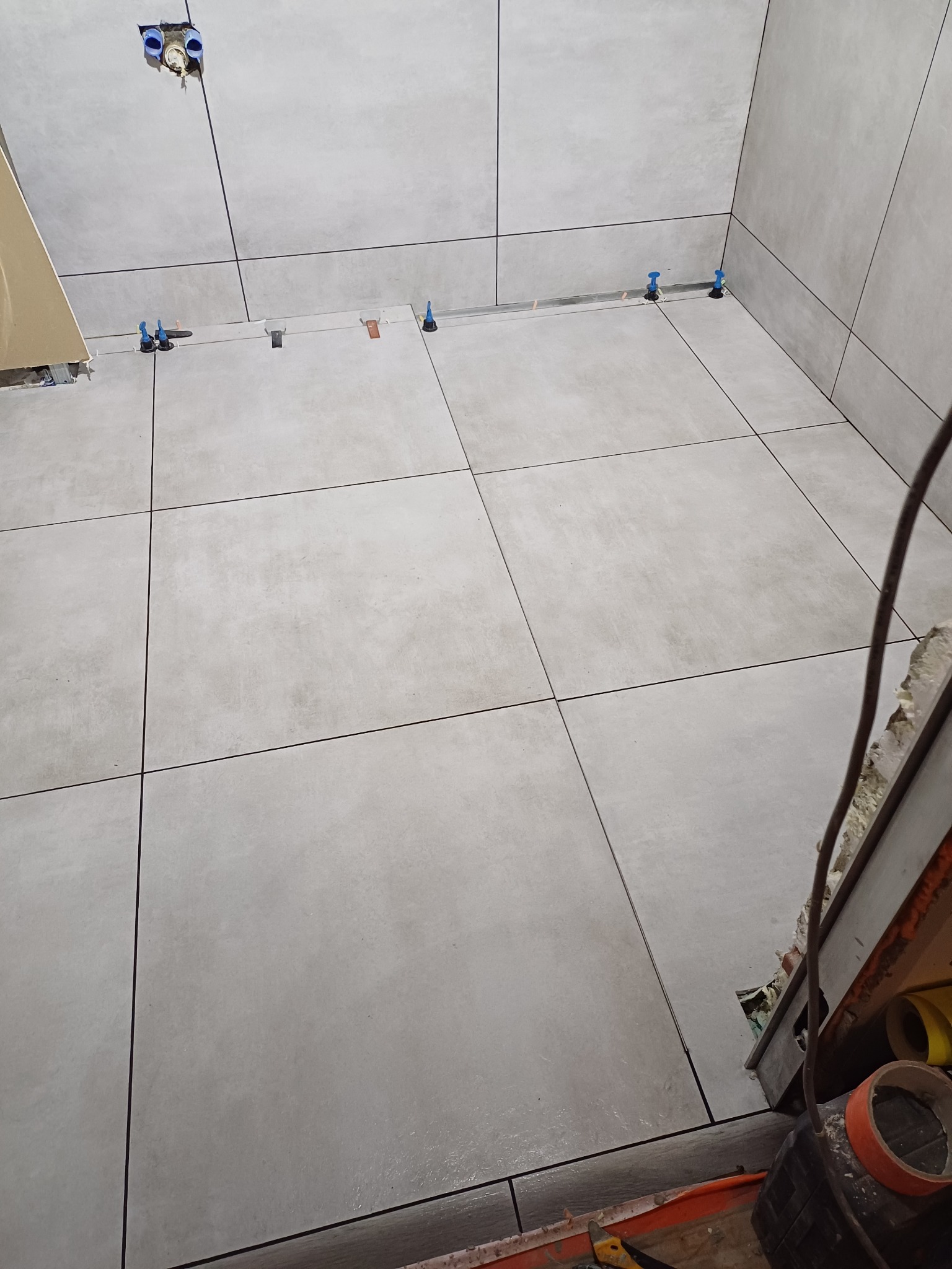 Świeżo ułożone, duże, szare płytki podłogowe i ścienne, z systemem poziomowania, widoczne instalacje wodne w ścianie, fragment narzędzi glazurniczych w rogu.