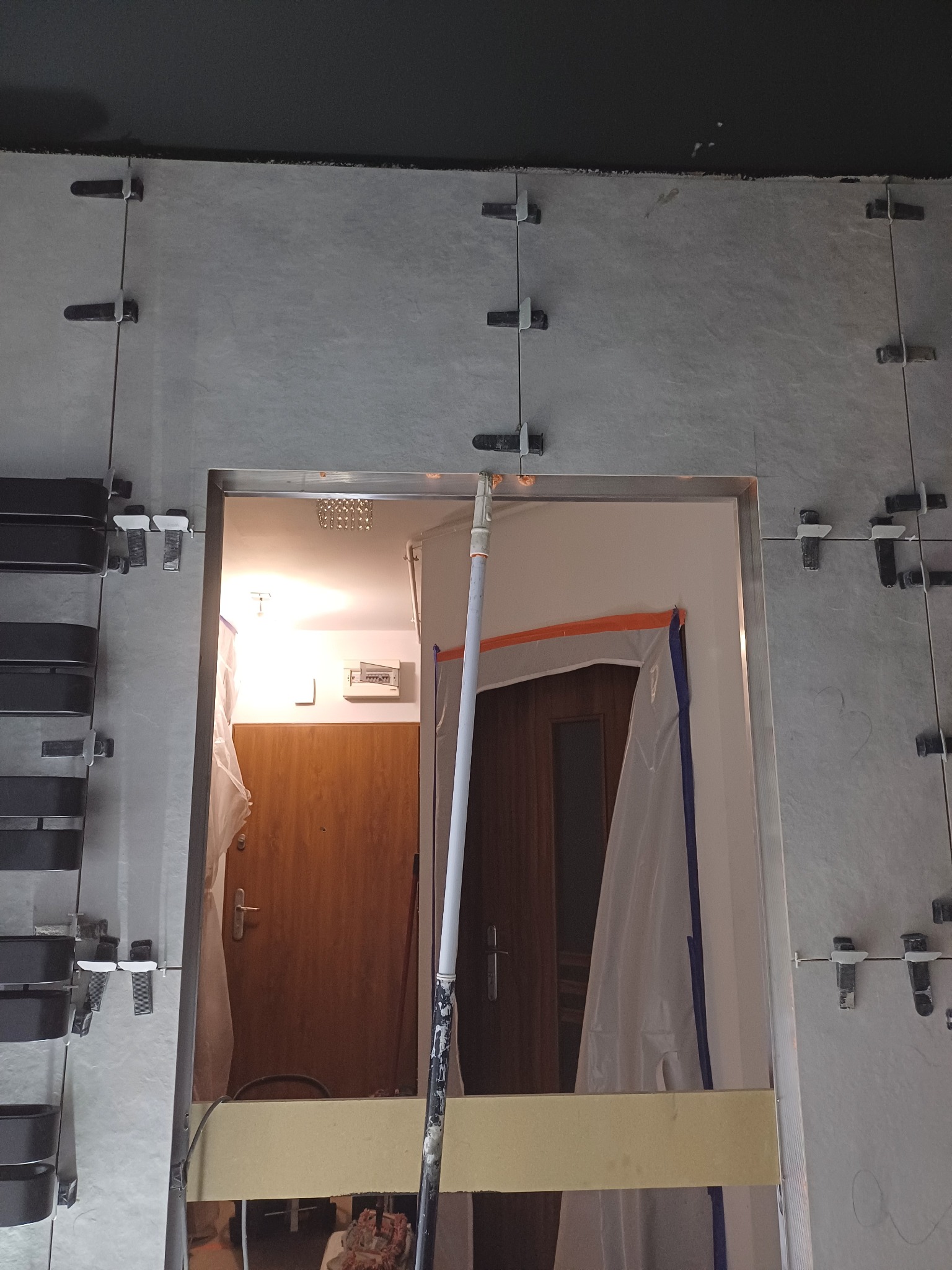 Świeżo ułożone szare płytki wokół futryny drzwi, zabezpieczone plastikowymi klipsami poziomującymi, widok na wnętrze mieszkania z drewnianymi drzwiami i osłoniętą folią framugą.