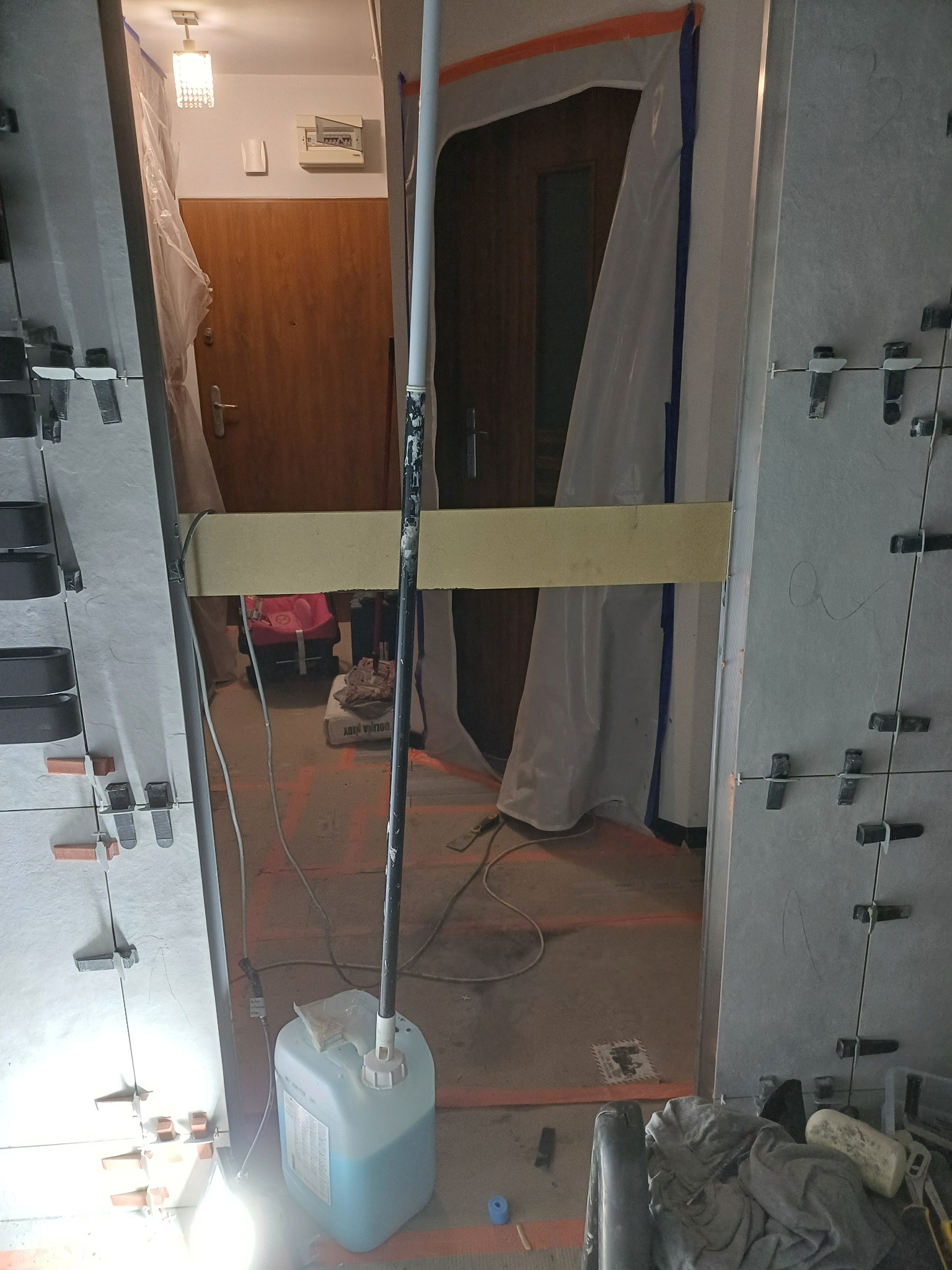 Świeżo ułożone szare płytki ścienne z krzyżykami dystansowymi, zabezpieczone folią drzwi wejściowe, wiadro z niebieskim płynem i narzędzia na podłodze.