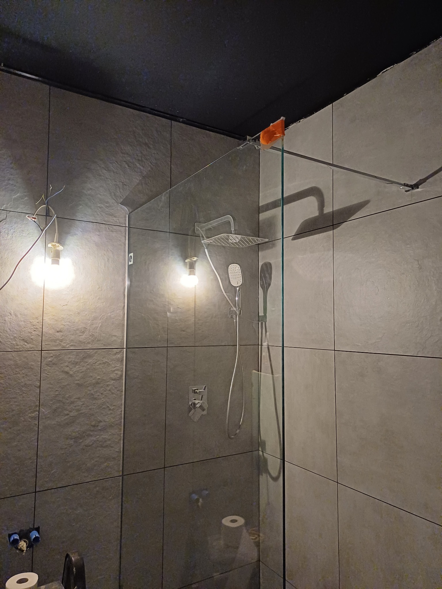 Szare płytki w łazience z zamontowaną szklaną kabiną prysznicową typu walk-in, widoczne podłączenia do baterii,  tymczasowe oświetlenie żarówkami i zabezpieczenie rogu kabiny pomarańczową folią.