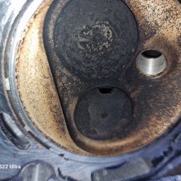 A.I.L Serwis - Detail of a dirty, used engine cylinder head with visible valves and threaded spark plug hole, showing signs of wear and carbon build-up.