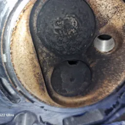 Detail of a dirty, used engine cylinder head with visible valves and threaded spark plug hole, showing signs of wear and carbon build-up.