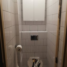 A.I.L Serwis - Wąska łazienka po remoncie z białymi płytkami, wiszącą szafką i zabudowaną spłuczką. Na desce sedesowej leżą narzędzia: młotek, poziomica i nożyk.