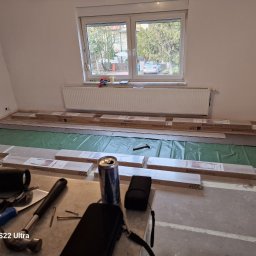 A.I.L Serwis - Rozpakowane panele podłogowe Kronostep na zielonej folii izolacyjnej, przygotowane do ułożenia w pokoju z grzejnikiem pod oknem, widoczne narzędzia i puszka napoju na pierwszym planie.