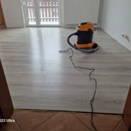 Szary panel podłogowy po umyciu, widoczny odkurzacz piorący w kolorach czarnym i pomarańczowym, kabel leżący na podłodze, fragment brązowej futryny i płytek podłogowych.