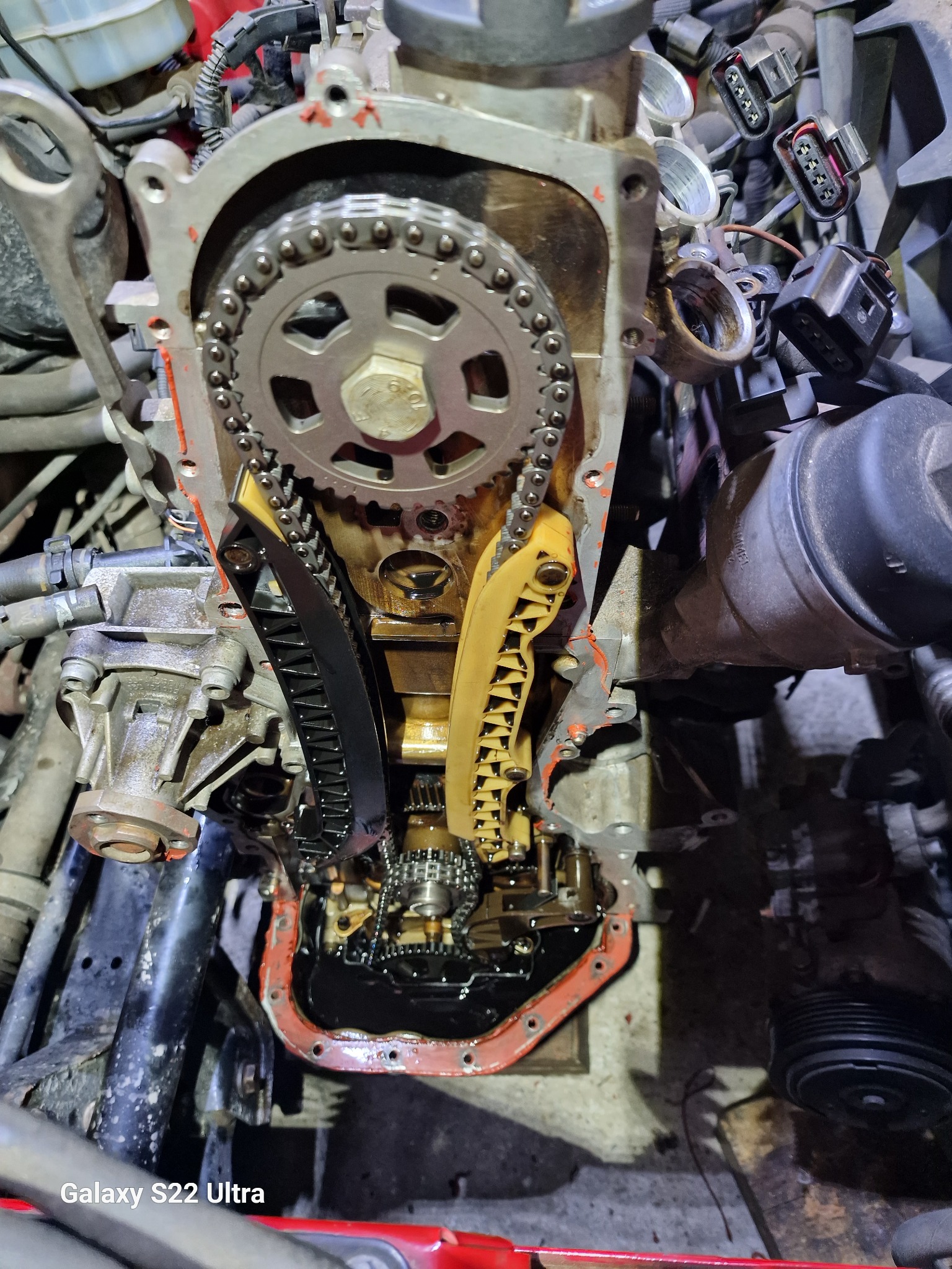 Otwarty silnik samochodowy z widocznym łańcuchem rozrządu, kołem zębatym i napinaczami łańcucha, zalany olejem.
