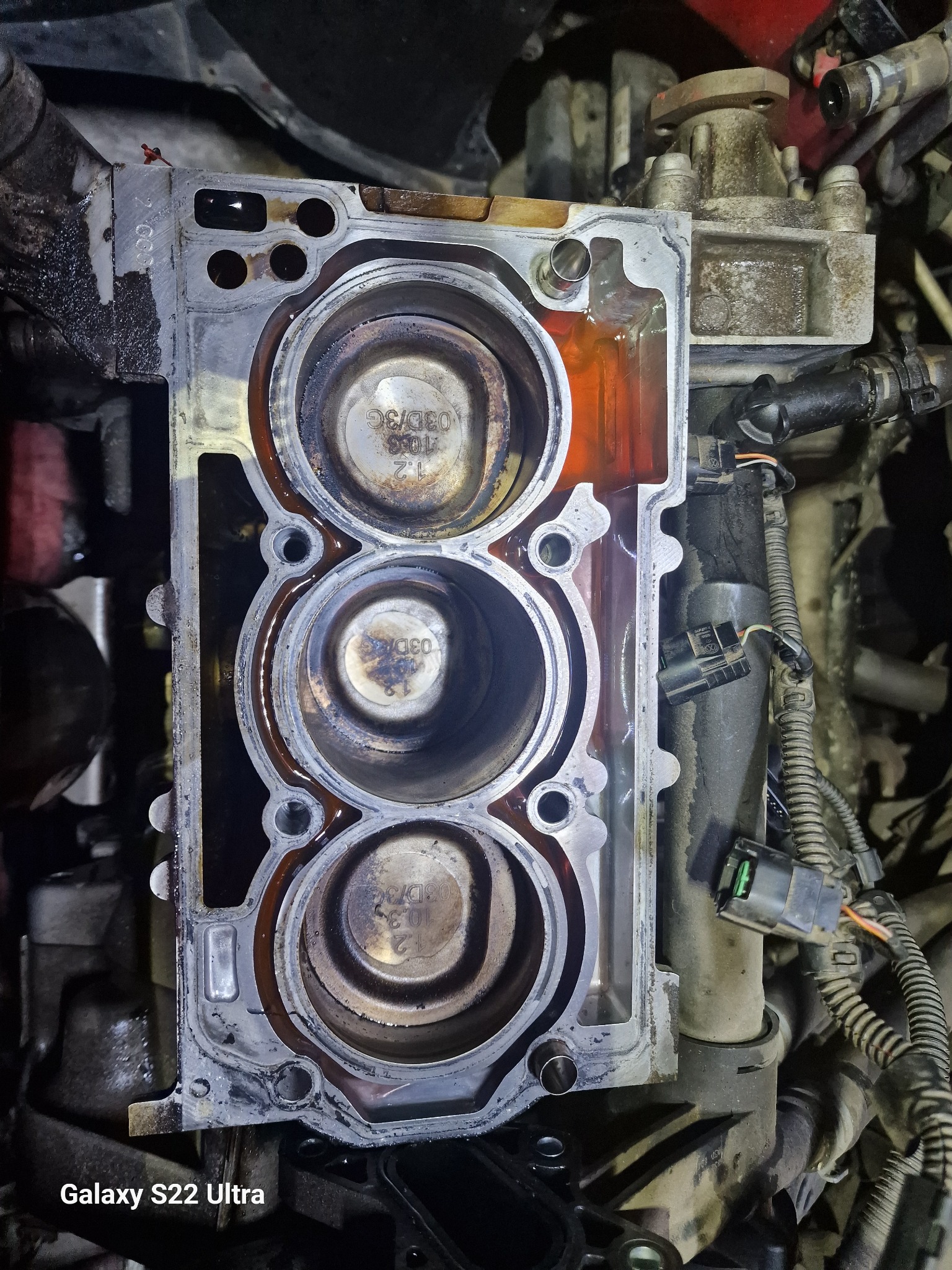 Otwarty blok silnika samochodowego z widocznymi trzema cylindrami, ślady oleju i nagaru, fragment instalacji elektrycznej w tle.