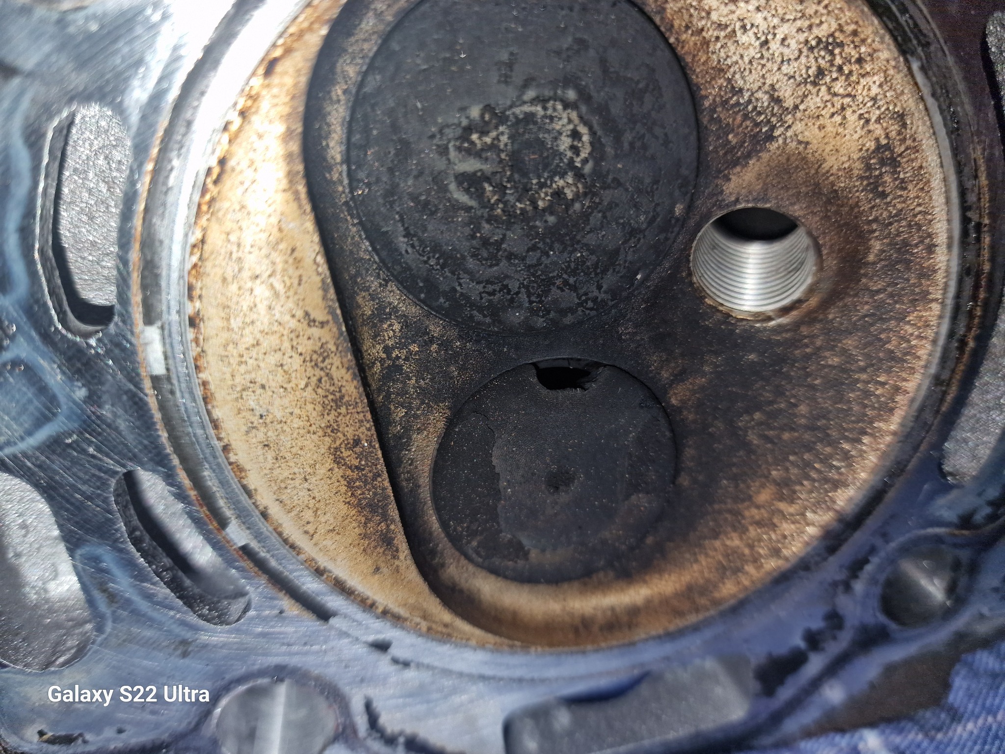 Detail of a dirty, used engine cylinder head with visible valves and threaded spark plug hole, showing signs of wear and carbon build-up.