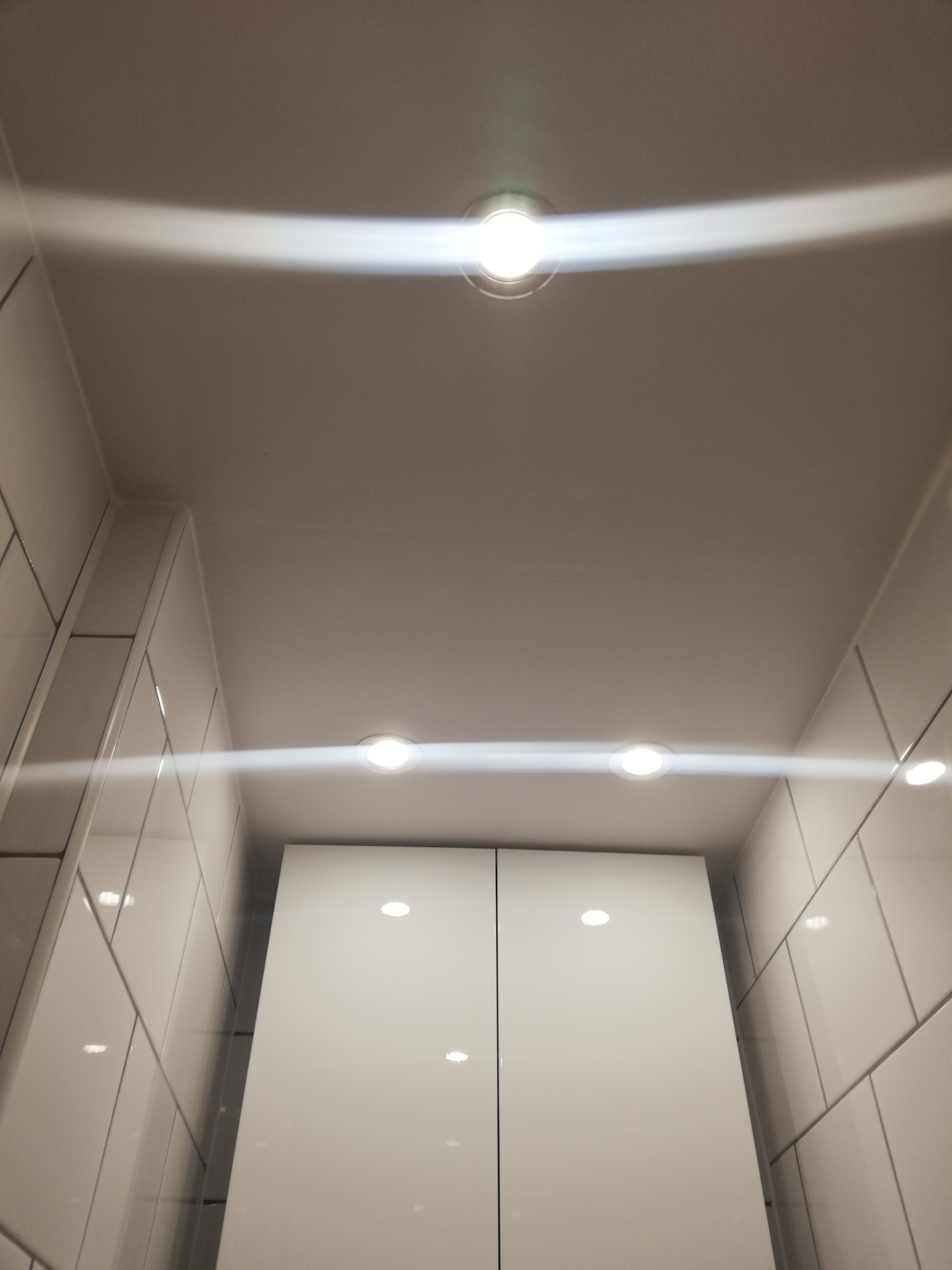 Sufit podwieszany z oświetleniem LED w łazience, białe płytki na ścianach i szafka z połyskiem.