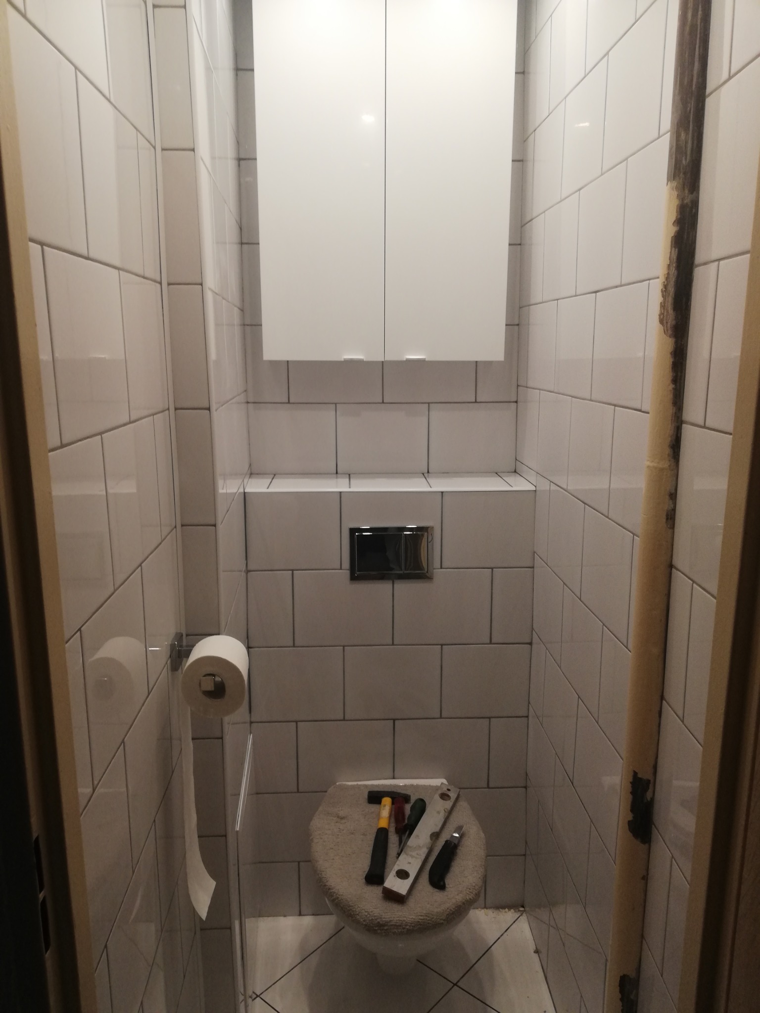 Wąska łazienka po remoncie z białymi płytkami, wiszącą szafką i zabudowaną spłuczką. Na desce sedesowej leżą narzędzia: młotek, poziomica i nożyk.