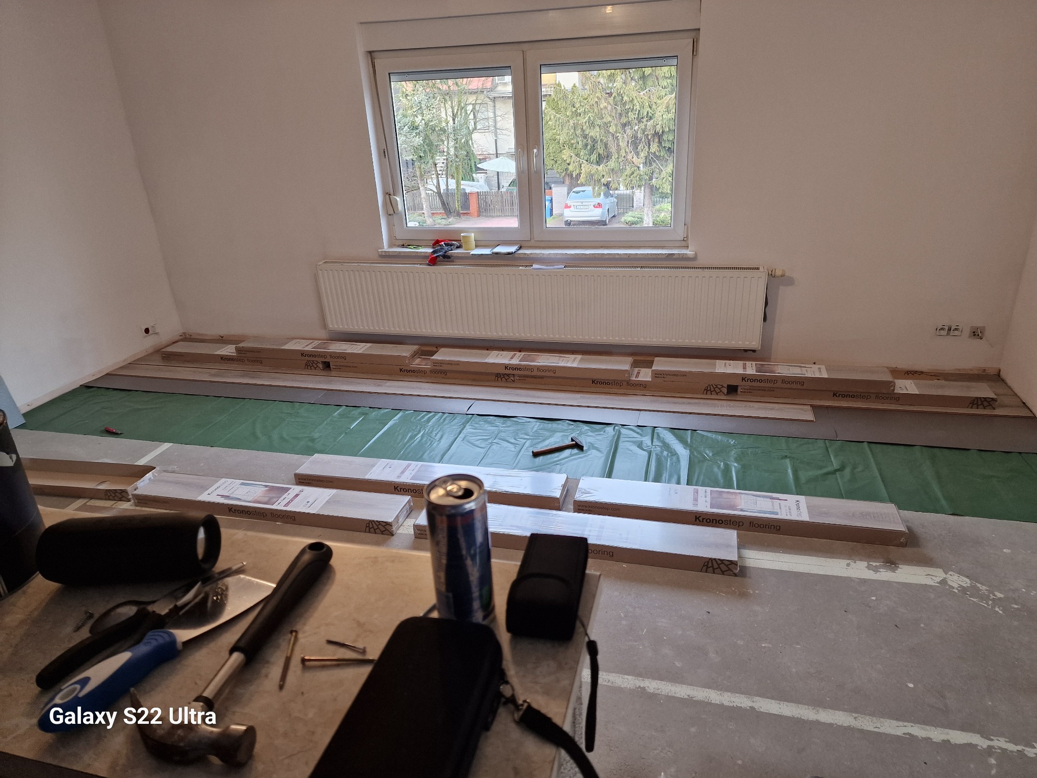 Rozpakowane panele podłogowe Kronostep na zielonej folii izolacyjnej, przygotowane do ułożenia w pokoju z grzejnikiem pod oknem, widoczne narzędzia i puszka napoju na pierwszym planie.