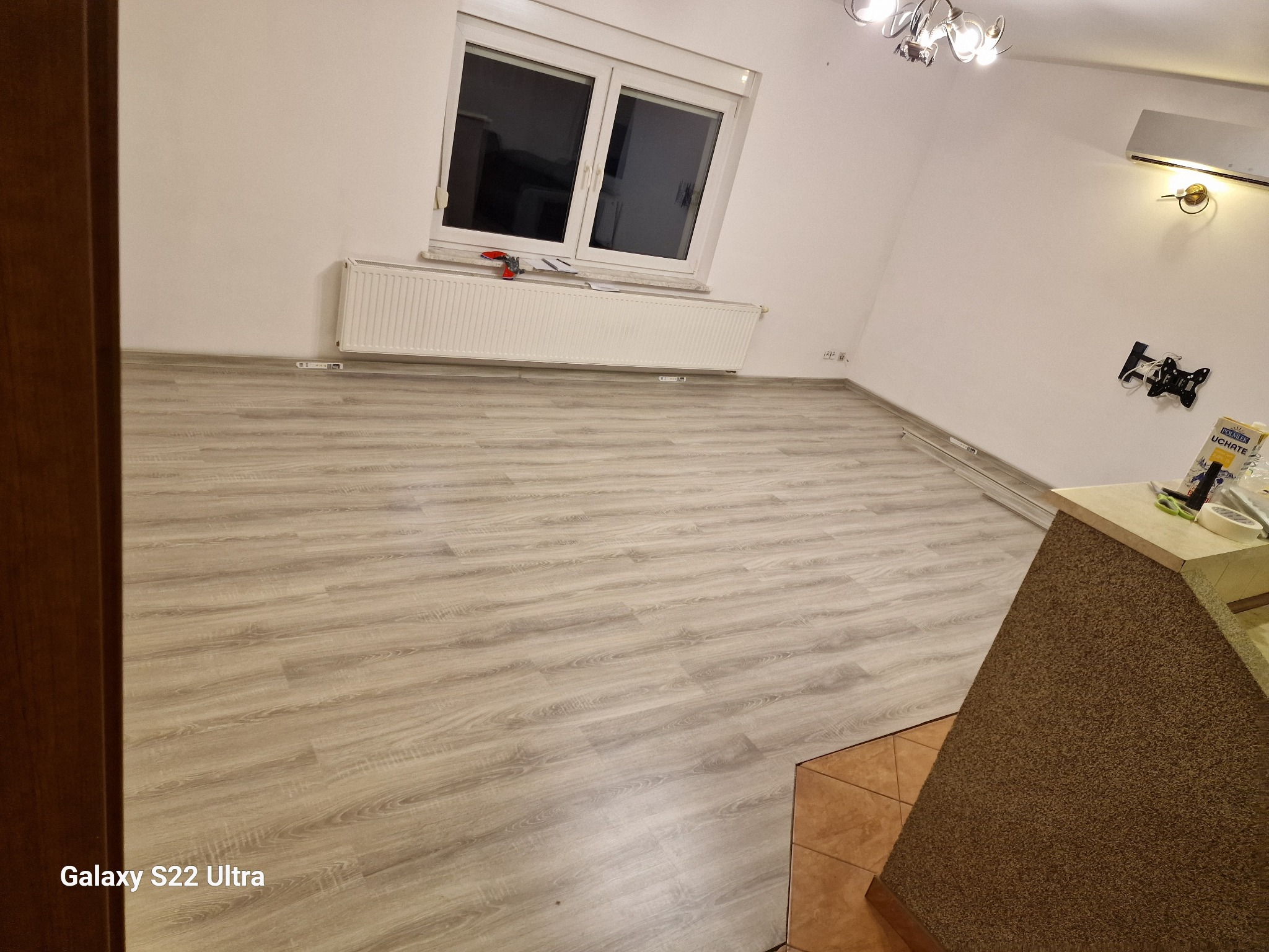 Jasne panele podłogowe w pokoju z białymi ścianami, grzejnikiem pod oknem i widocznym fragmentem płytek podłogowych w przejściu do innego pomieszczenia.