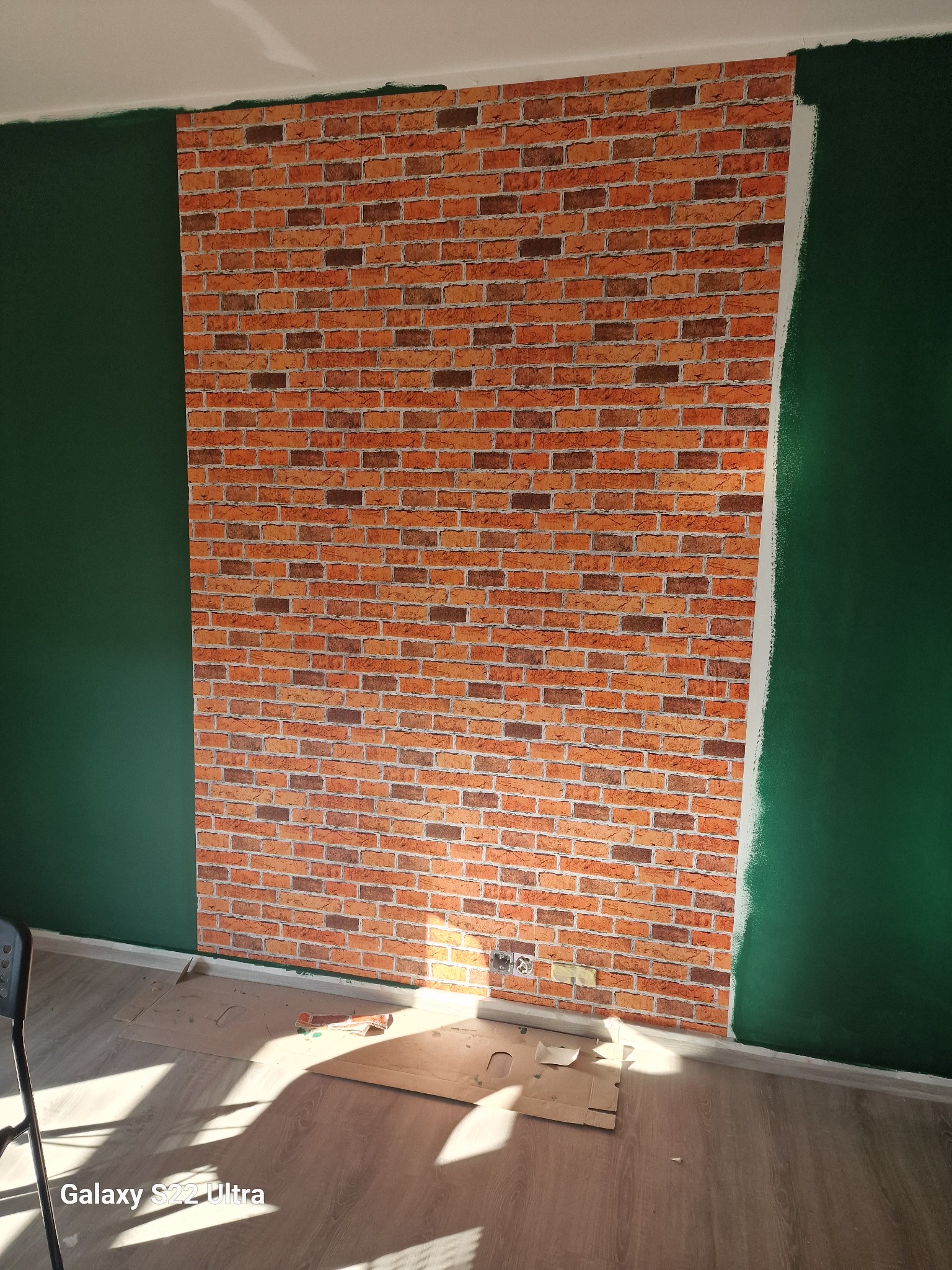 Ściana z tapetą imitującą cegłę, sąsiadująca z pomalowaną na zielono ścianą, widoczne fragmenty podłogi i kartonu, w trakcie prac wykończeniowych.