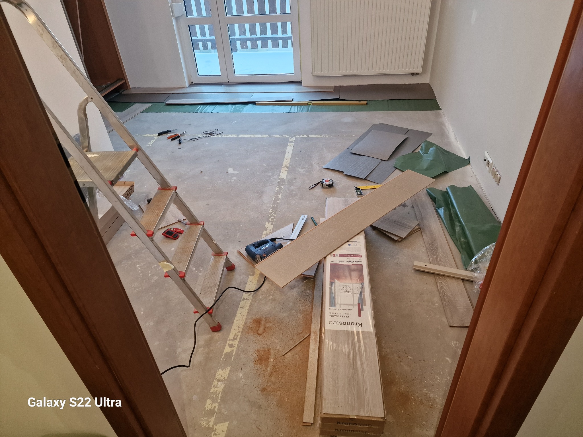 Przygotowania do układania paneli podłogowych: rozłożone podkłady izolacyjne, paczka paneli Kronostep, wyrzynarka, miara i poziomica na podłodze, drabina oparta o ścianę, widok zza futryny.