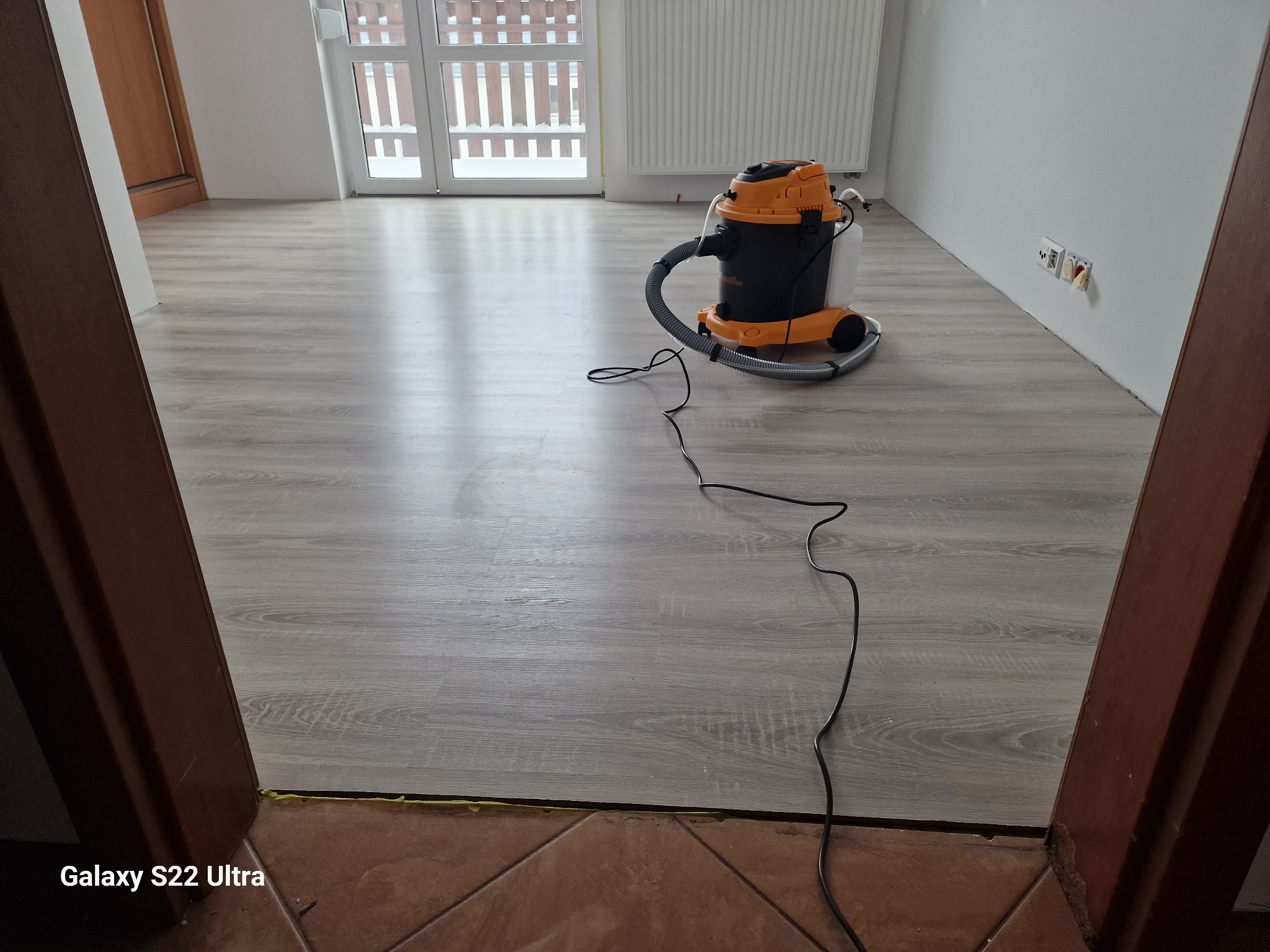 Szary panel podłogowy po umyciu, widoczny odkurzacz piorący w kolorach czarnym i pomarańczowym, kabel leżący na podłodze, fragment brązowej futryny i płytek podłogowych.