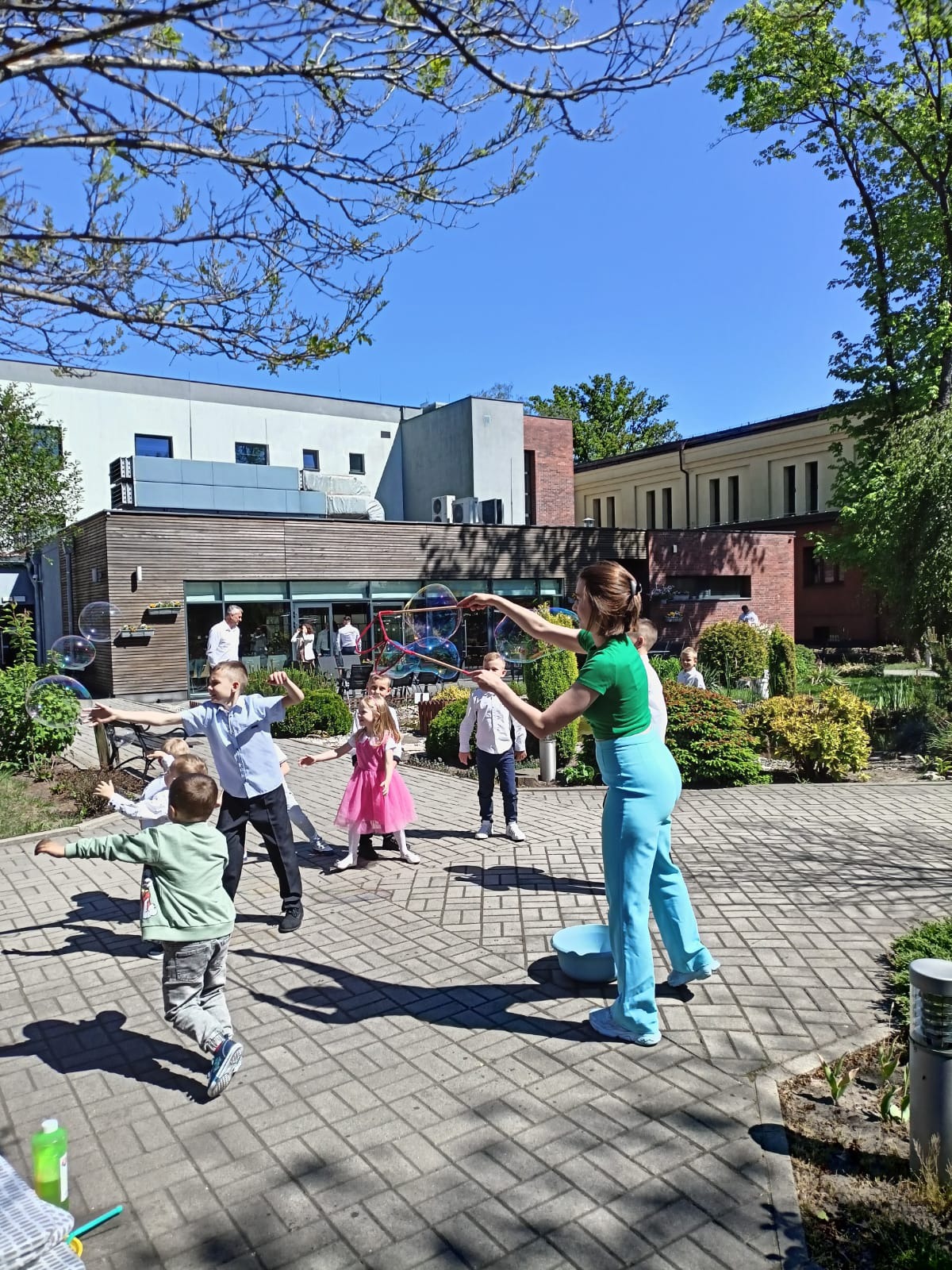 Animator w zielonej koszulce puszcza ogromne bańki mydlane dla grupy dzieci na brukowanym placu przed budynkiem w słoneczny dzień. Radosna atmosfera.