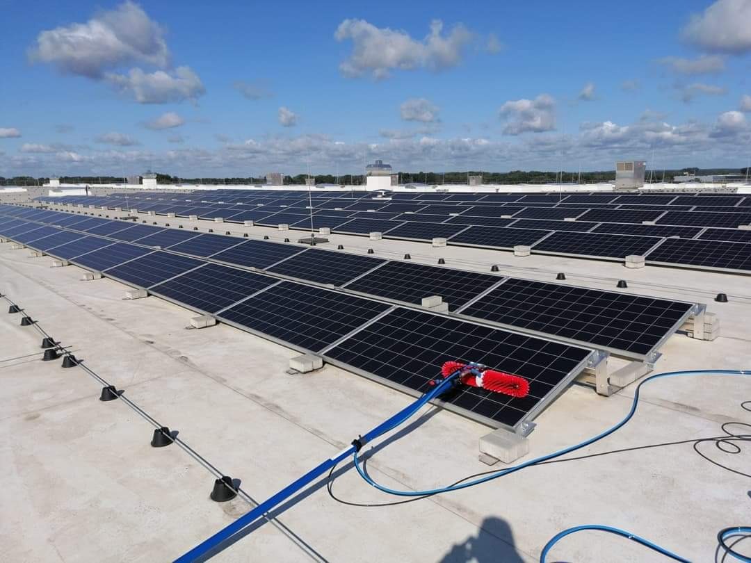 Czyszczenie paneli słonecznych na dachu budynku przemysłowego za pomocą szczotki z doprowadzeniem wody pod ciśnieniem, błękitne niebo z chmurami w tle.