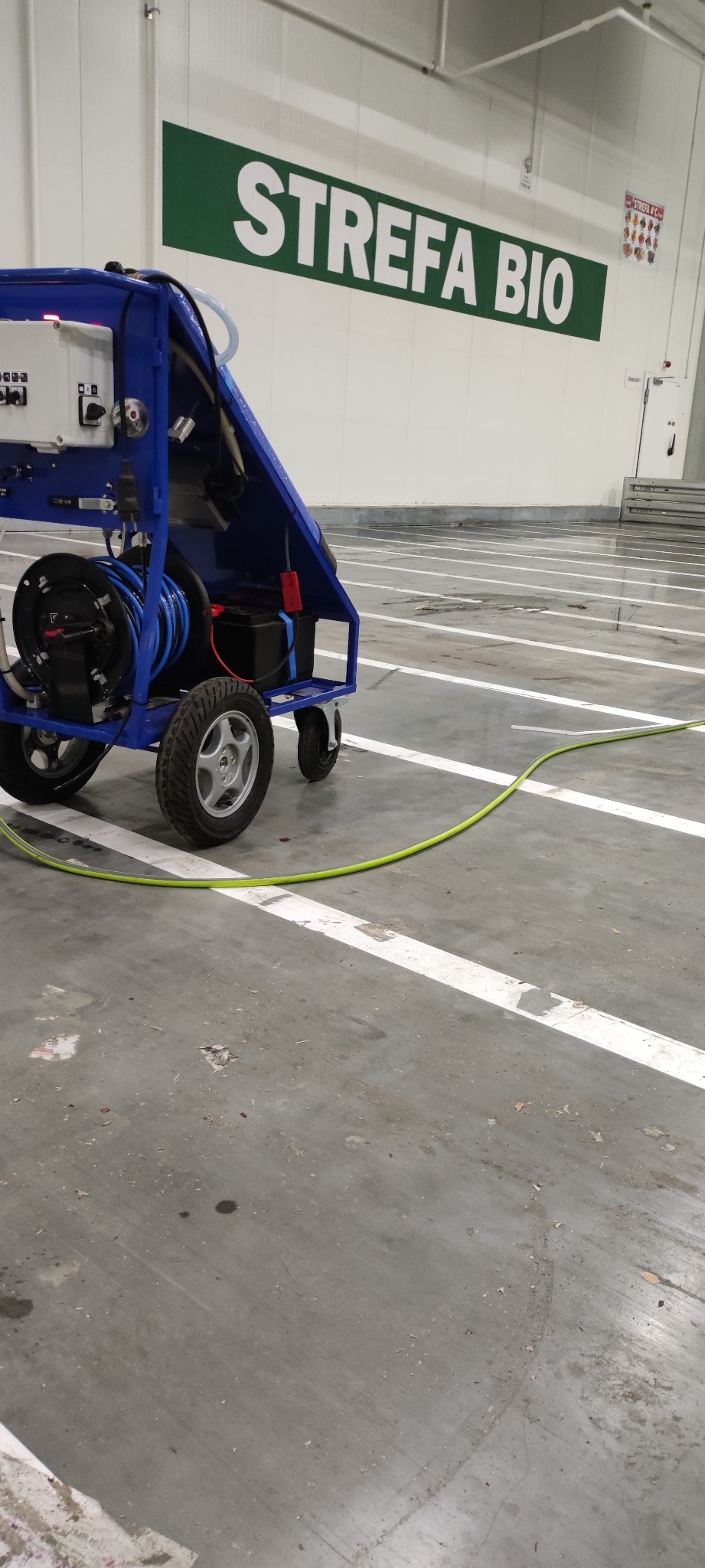 Wózek z urządzeniem wysokociśnieniowym do czyszczenia przemysłowego na hali produkcyjnej, widoczny napis 'STREFA BIO' na ścianie w tle.