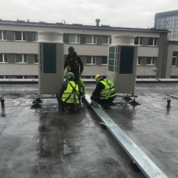 JAGODZIK MAREK ACC KLIMAR KLIMATYZACJA, WENTYLACJA CHŁODNICTWO - Trzech monterów w odblaskowych kamizelkach instaluje klimatyzatory na dachu budynku, widoczne metalowe koryta kablowe, pochmurne niebo.
