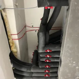 Rzut z dołu na izolowane rury wentylacyjne z czerwonymi zaworami, prowadzone pod sufitem, z widoczną izolacją wełną mineralną i przewodami elektrycznymi.