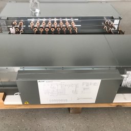 JAGODZIK MAREK ACC KLIMAR KLIMATYZACJA, WENTYLACJA CHŁODNICTWO - Dwa moduły klimatyzacyjne Mitsubishi Electric, jeszcze zapakowane, leżą na drewnianej palecie. Widoczne miedziane rurki przyłączeniowe i schemat elektryczny na obudowie.