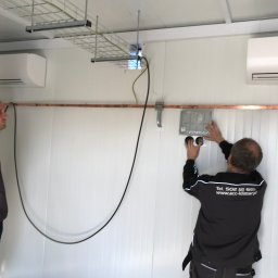 JAGODZIK MAREK ACC KLIMAR KLIMATYZACJA, WENTYLACJA CHŁODNICTWO - Instalacja klimatyzacji w pomieszczeniu: monter w czarnej bluzie z logo firmy montuje panel sterowania na ścianie, widoczne dwie jednostki wewnętrzne klimatyzacji oraz kable w korytku.