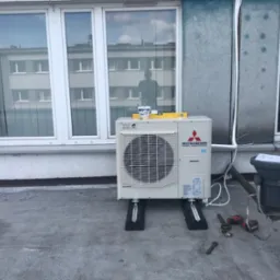 Zewnętrzna jednostka klimatyzacji Mitsubishi Heavy Industries zamontowana na dachu budynku z widocznymi narzędziami monterskimi, poziomnicą i kubkiem z narzędziami, odbicie sylwetki osoby w oknie.