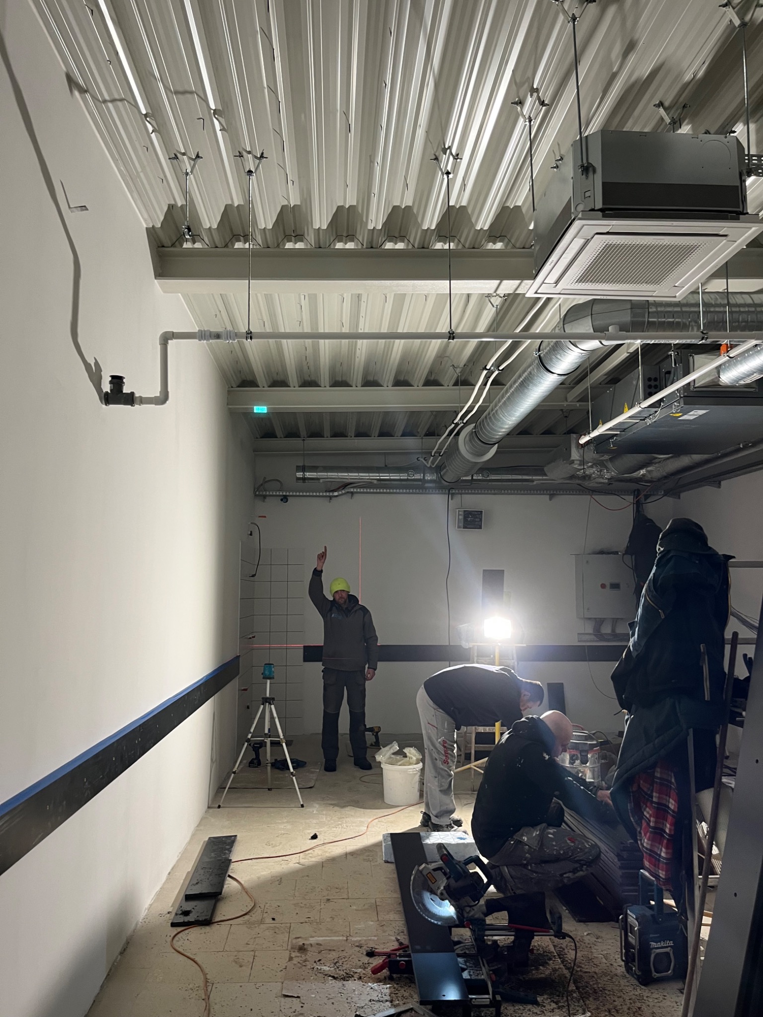 Montaż systemu wentylacji w obiekcie komercyjnym: trzech instalatorów pracuje przy układaniu kanałów wentylacyjnych pod sufitem, jeden z nich używa lasera do precyzyjnego pomiaru.