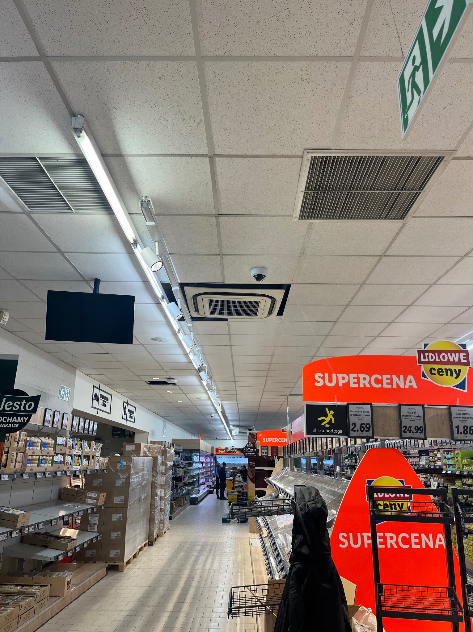 Sufit w supermarkecie z widocznymi elementami wentylacji i klimatyzacji: kratki wentylacyjne, czujnik oraz oświetlenie liniowe.