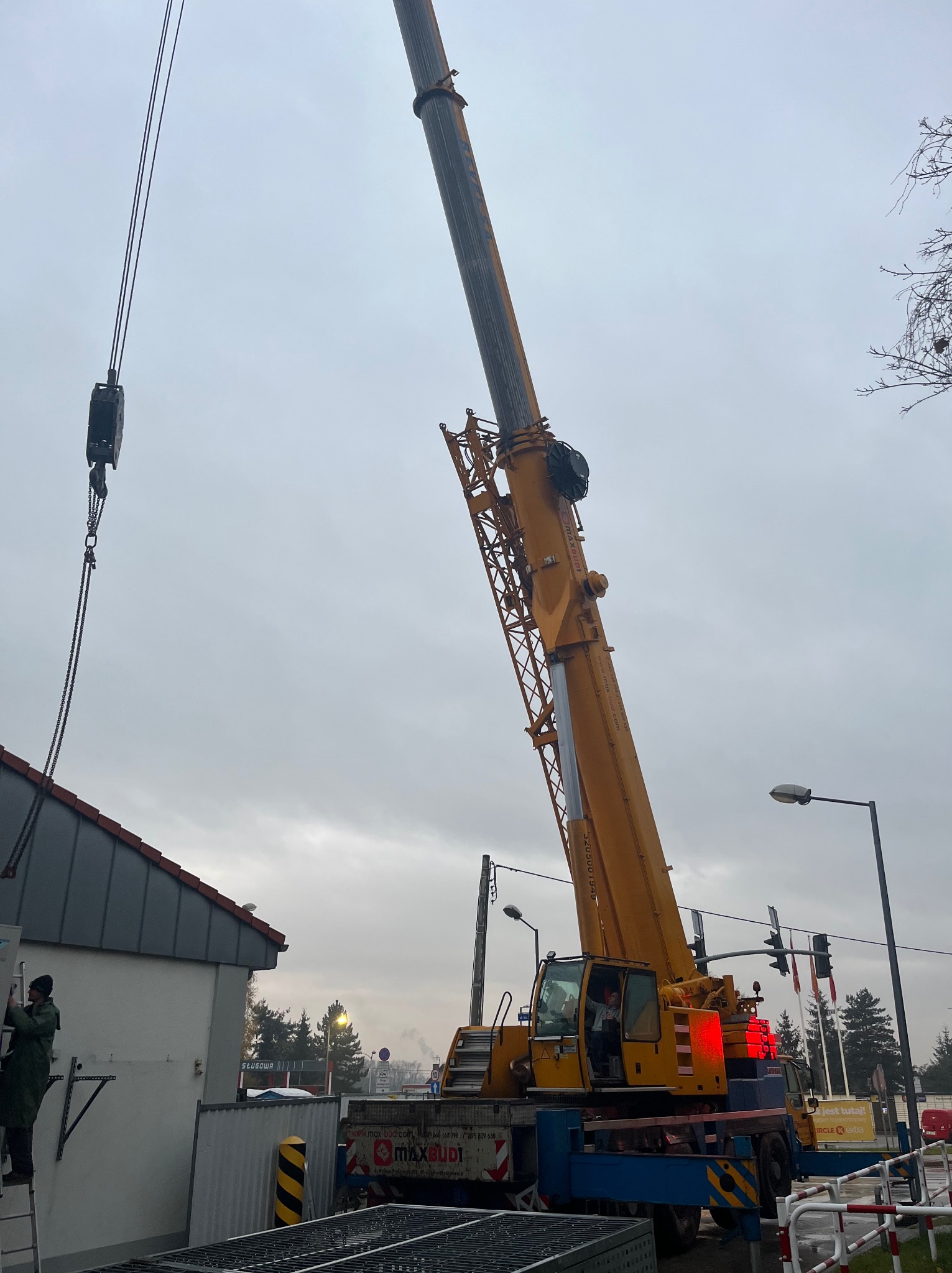 Montaż klimatyzacji zewnętrznej na budynku, z użyciem żółtego dźwigu Maxbud, pracownik na drabinie przy jednostce, widoczne elementy instalacji i sygnalizacja świetlna w tle.
