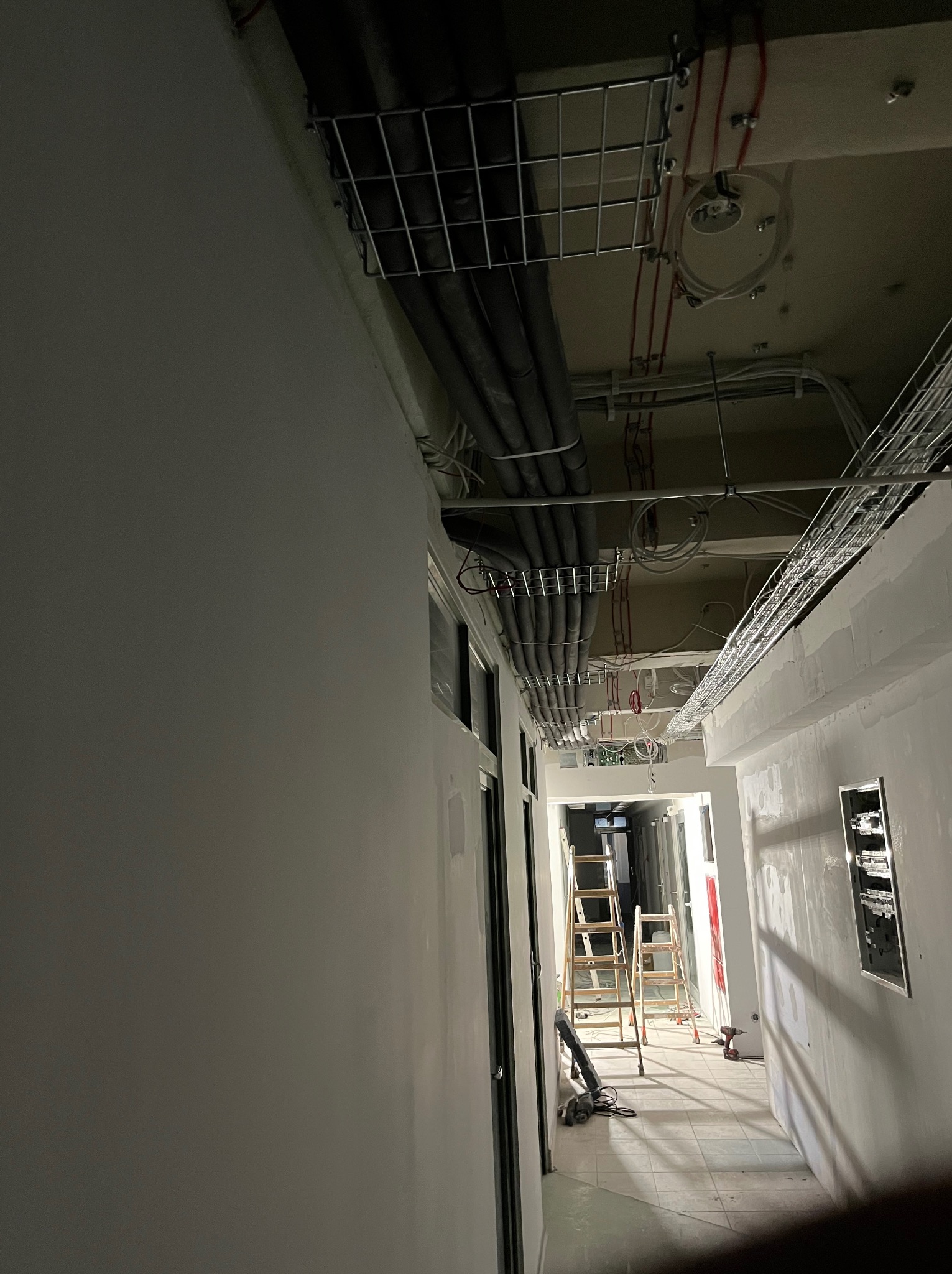 Korytarz w trakcie montażu instalacji wentylacyjnych pod sufitem, widoczne koryta kablowe, rury izolowane i drabiny monterskie.