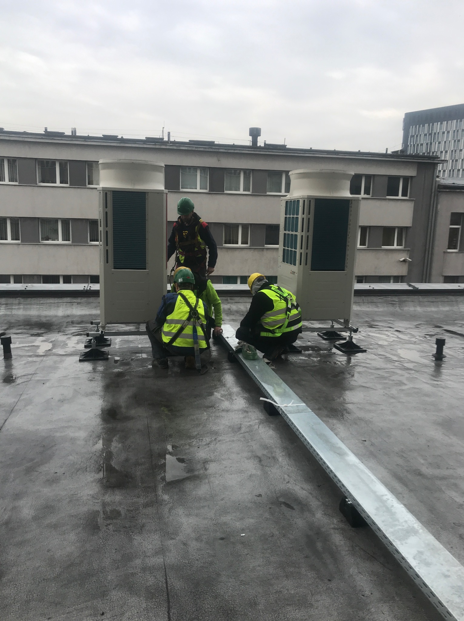 Trzech monterów w odblaskowych kamizelkach instaluje klimatyzatory na dachu budynku, widoczne metalowe koryta kablowe, pochmurne niebo.