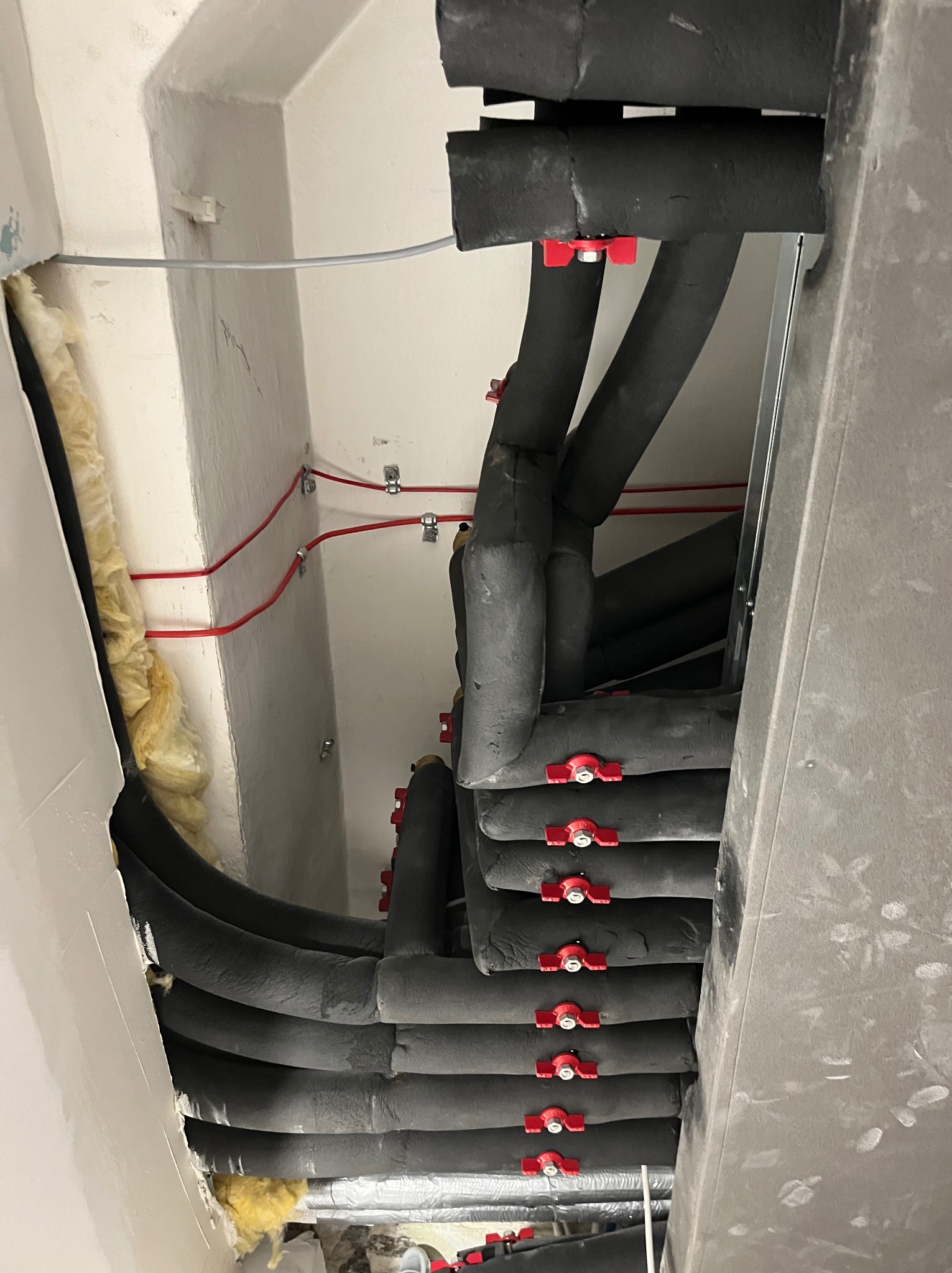 Rzut z dołu na izolowane rury wentylacyjne z czerwonymi zaworami, prowadzone pod sufitem, z widoczną izolacją wełną mineralną i przewodami elektrycznymi.