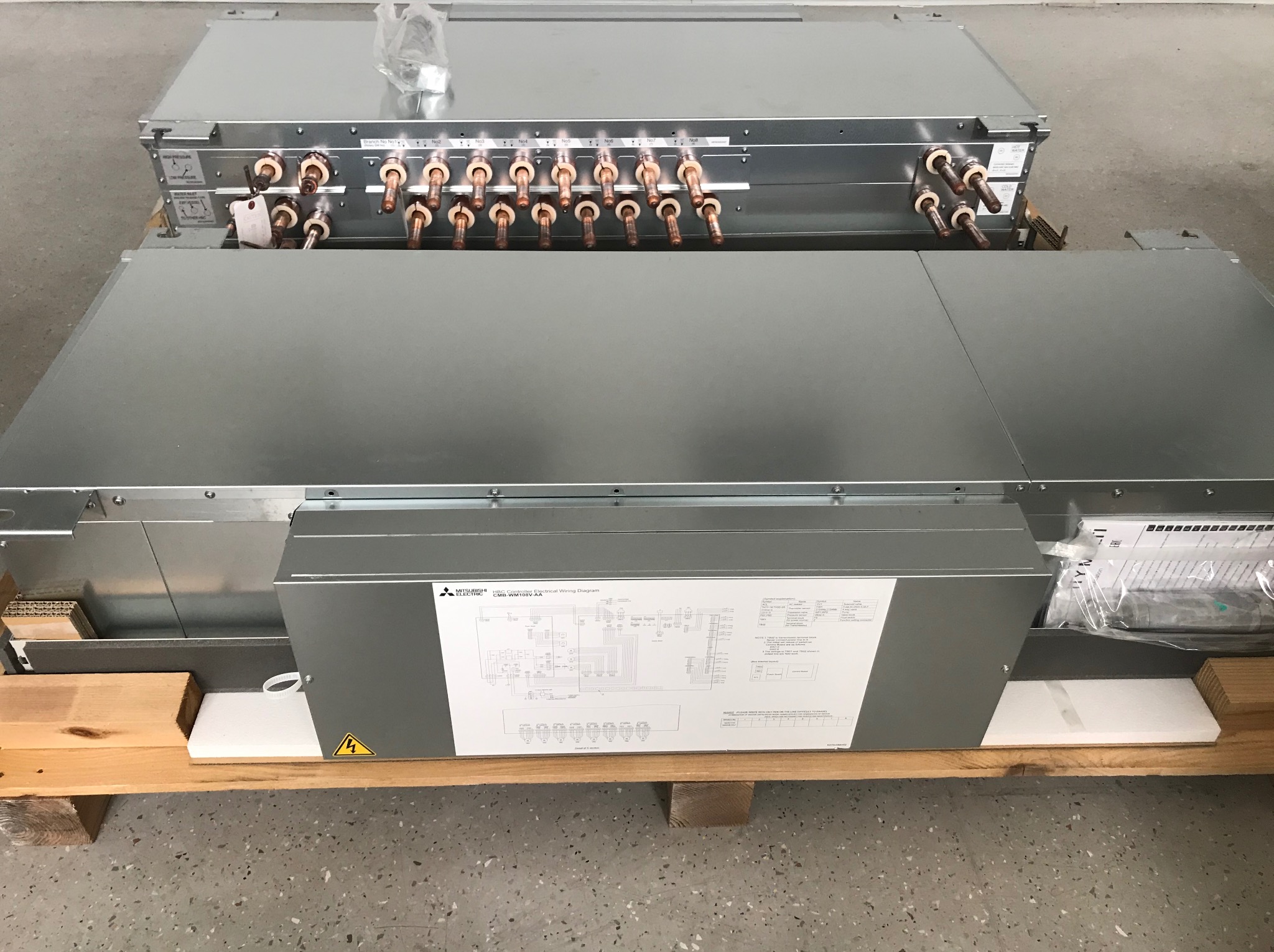 Dwa moduły klimatyzacyjne Mitsubishi Electric, jeszcze zapakowane, leżą na drewnianej palecie. Widoczne miedziane rurki przyłączeniowe i schemat elektryczny na obudowie.