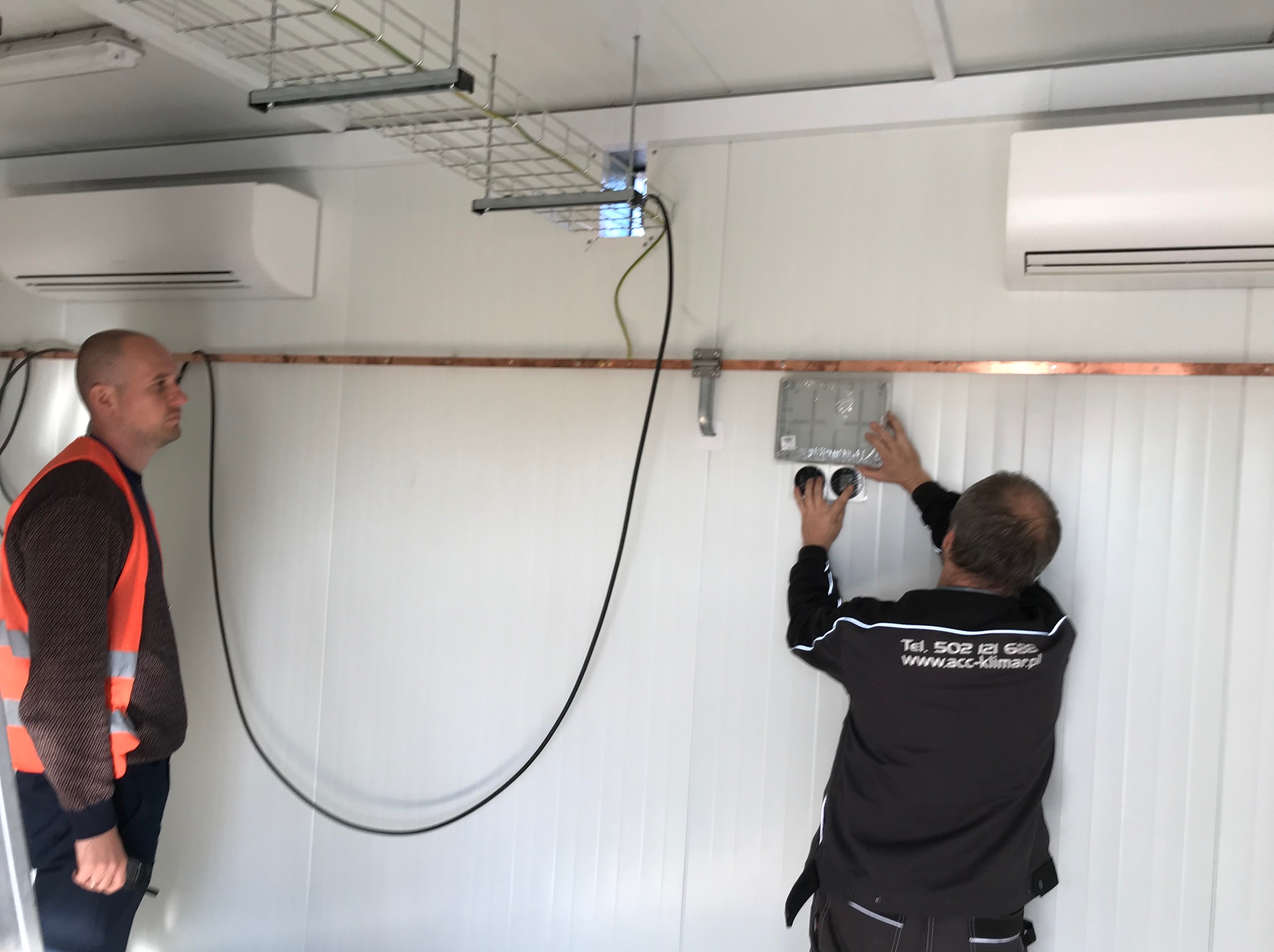 Instalacja klimatyzacji w pomieszczeniu: monter w czarnej bluzie z logo firmy montuje panel sterowania na ścianie, widoczne dwie jednostki wewnętrzne klimatyzacji oraz kable w korytku.