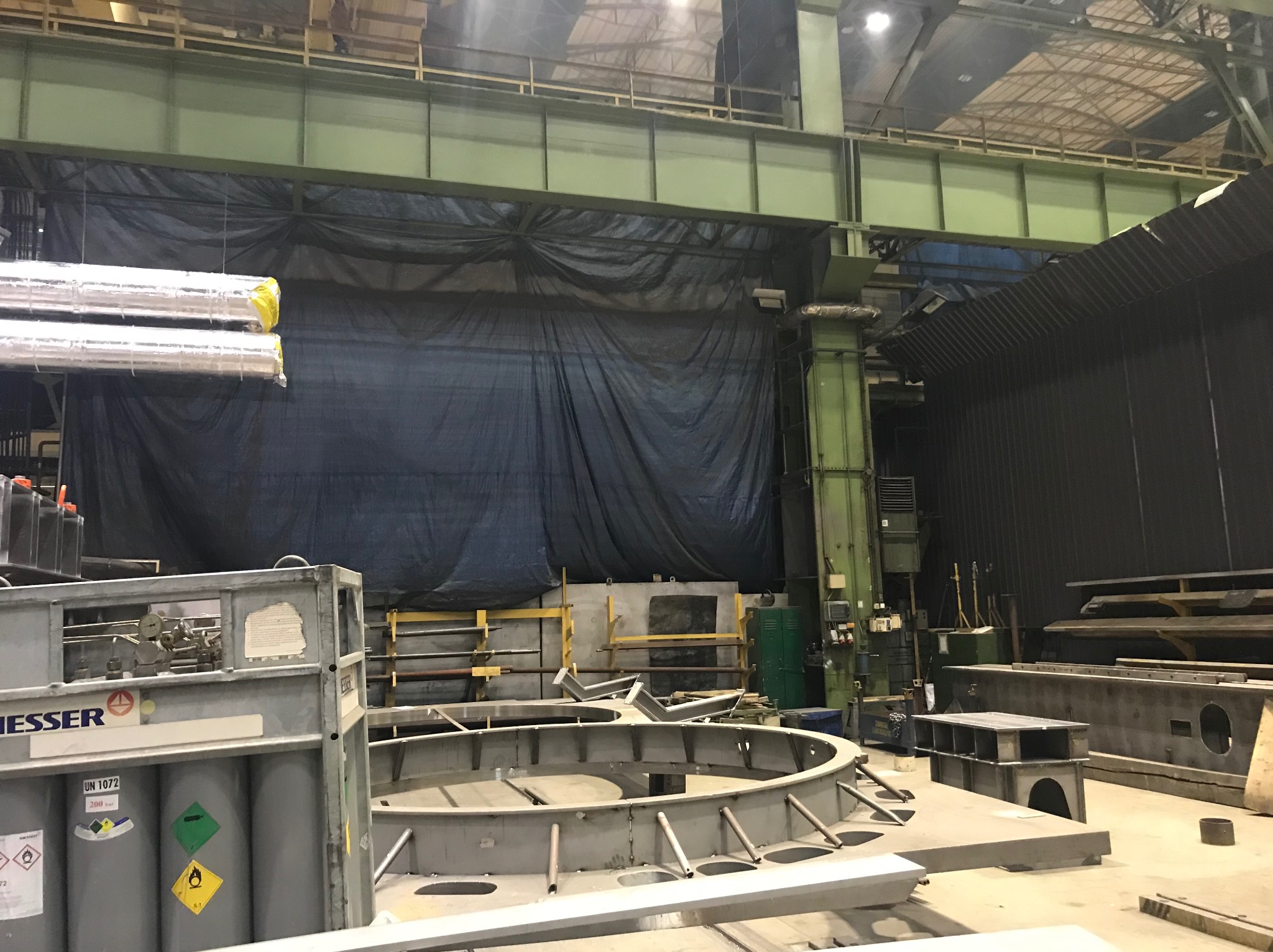 Wnętrze hali przemysłowej z konstrukcjami stalowymi, zbiornikami gazów technicznych Messer i rurami owiniętymi folią, oświetlone górnym światłem.