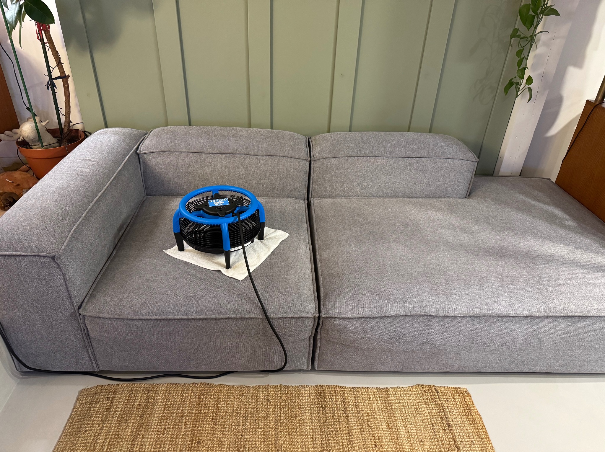 Szara, modułowa kanapa z niebiesko-czarnym wentylatorem do osuszania na białej ściereczce; kabel wentylatora biegnie wzdłuż kanapy na tle plecionej słomianki.