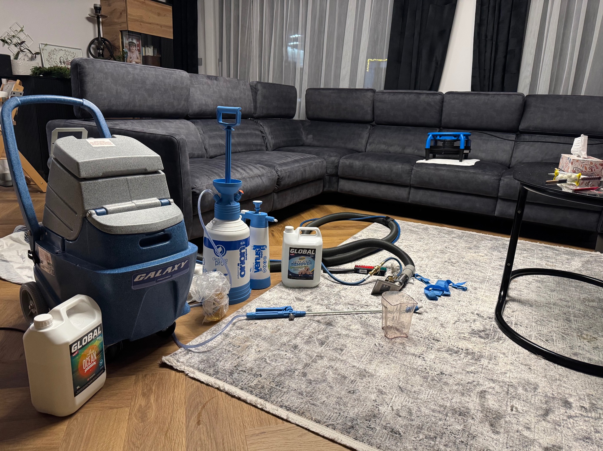 Sprzęt do czyszczenia dywanów i kanap, w tym odkurzacz piorący Galaxy Prochem, środki czyszczące Global, węże i końcówki, rozłożony na dywanie w salonie z szarą narożną sofą w tle.